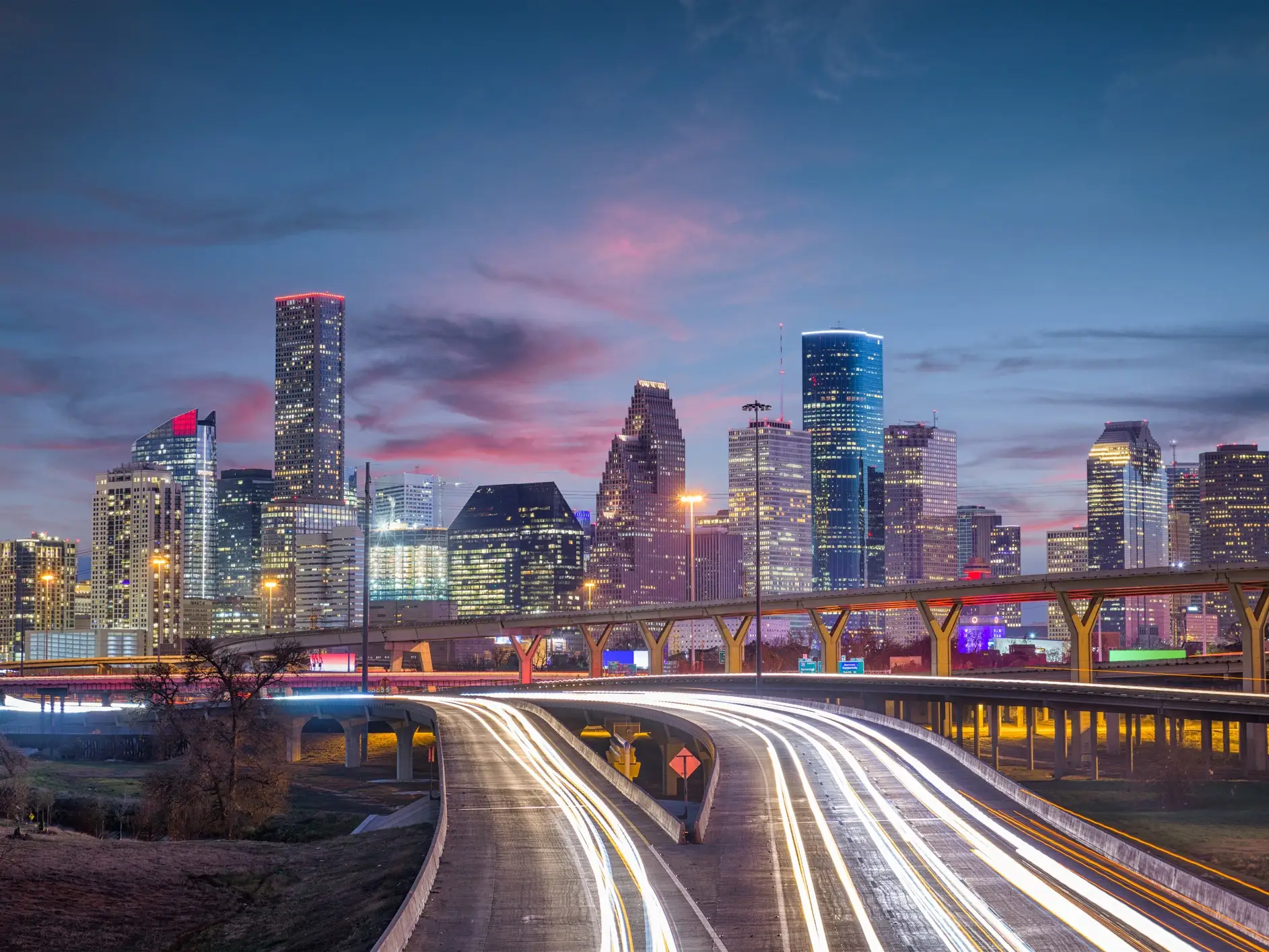 Downtown Houston skyline rises behind illuminated freeways and light trails at dusk.