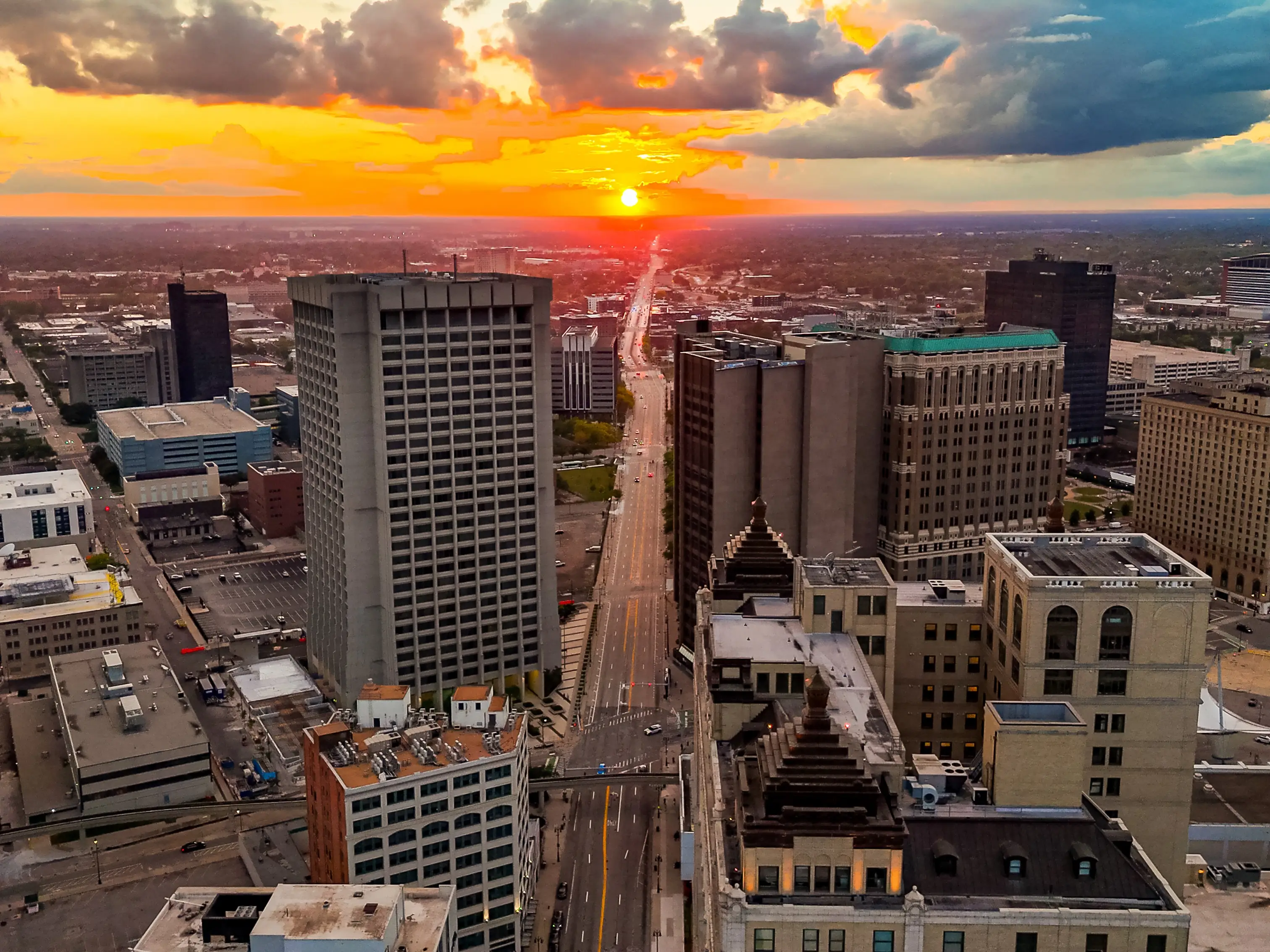 Downtown high-rises and a long central avenue stretch toward a bright sunset under dramatic clouds.