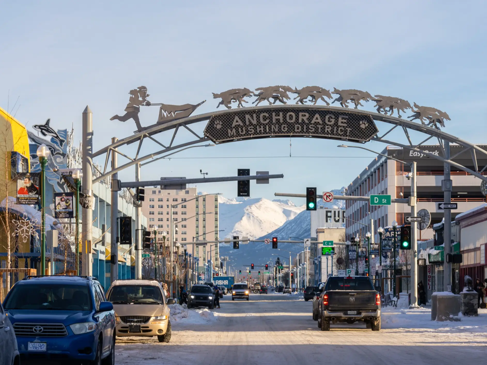 Snowy downtown Anchorage street framed by the Anchorage Mushing District arch with mountains visible in the distance.
