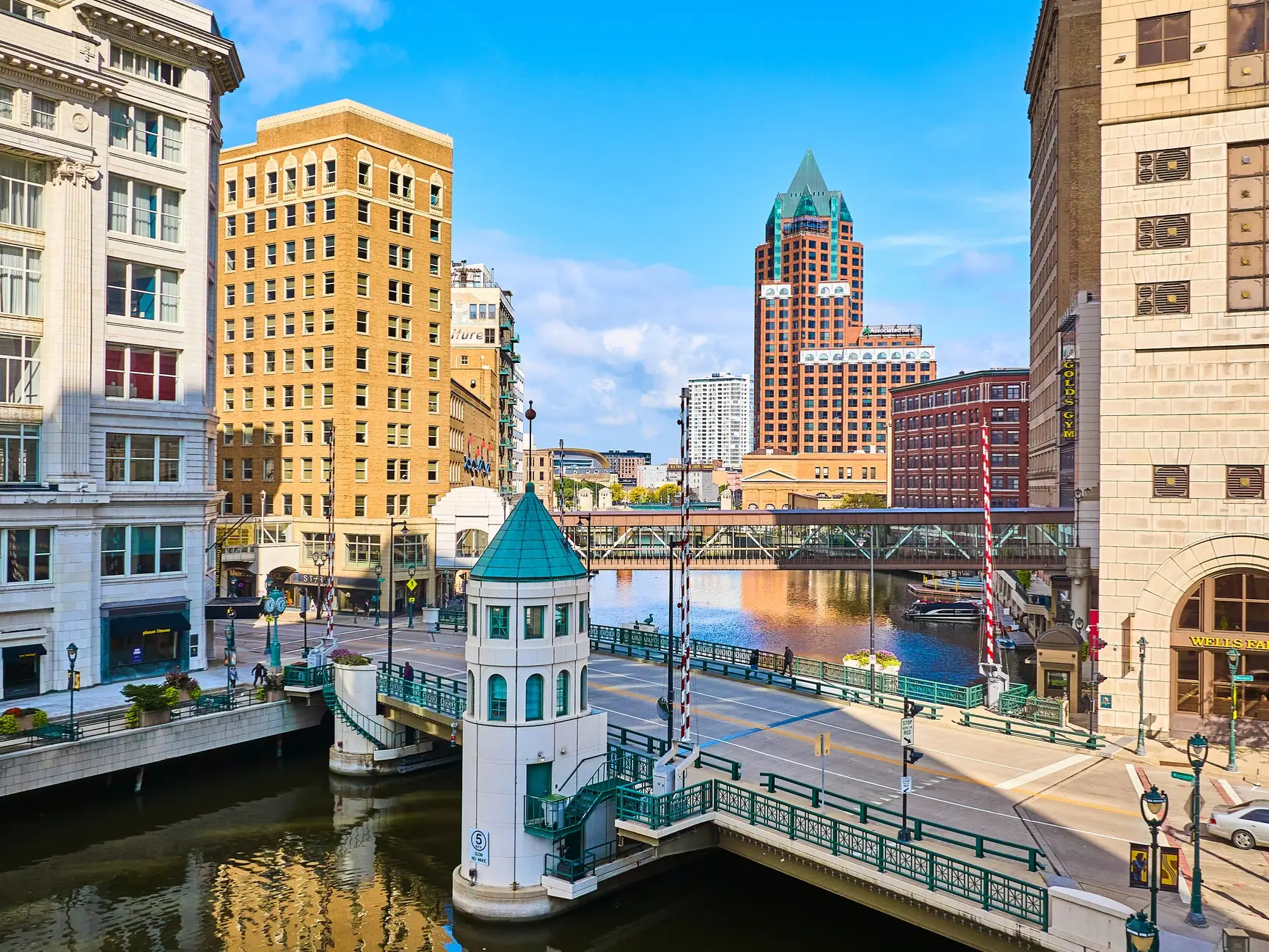 Downtown Milwaukee riverfront with bridges, skywalks, historic buildings, and a green-roofed bridge tender tower.