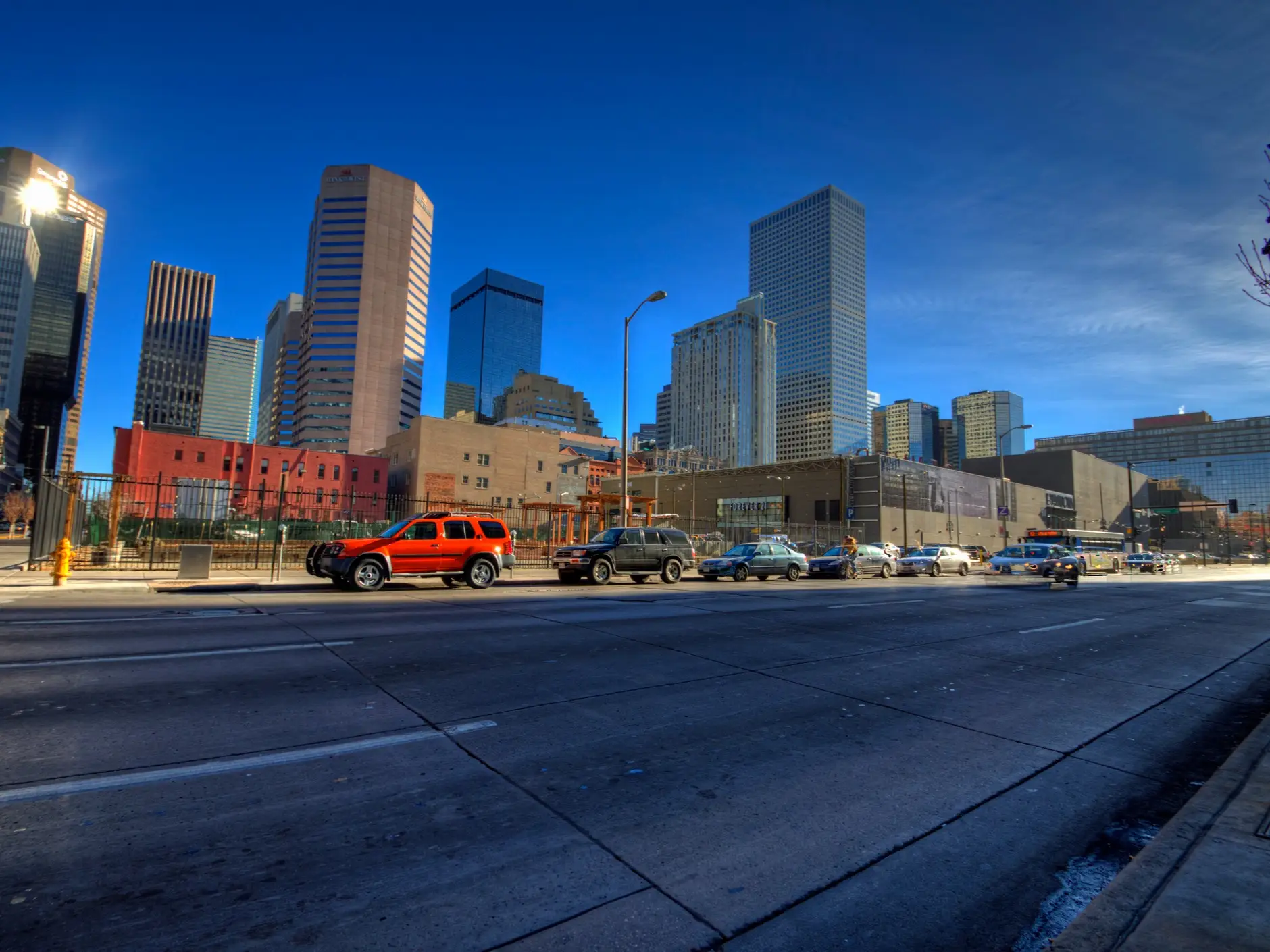 Cars line a downtown Denver street below high-rise buildings and the Denver Pavilions under a blue sky.