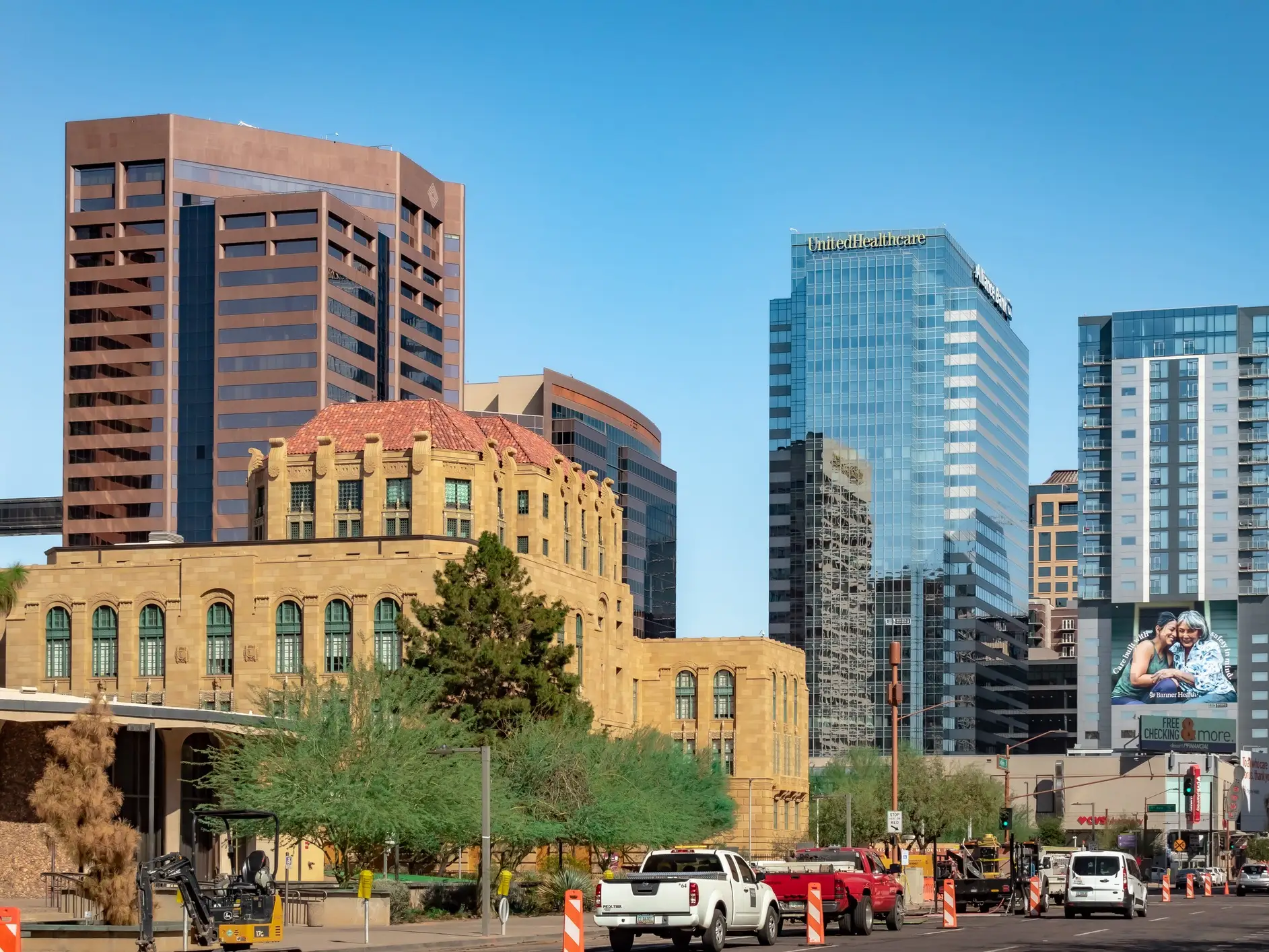 Downtown street with construction barriers, vehicles, a historic tan building, and high-rises including a UnitedHealthcare tower.