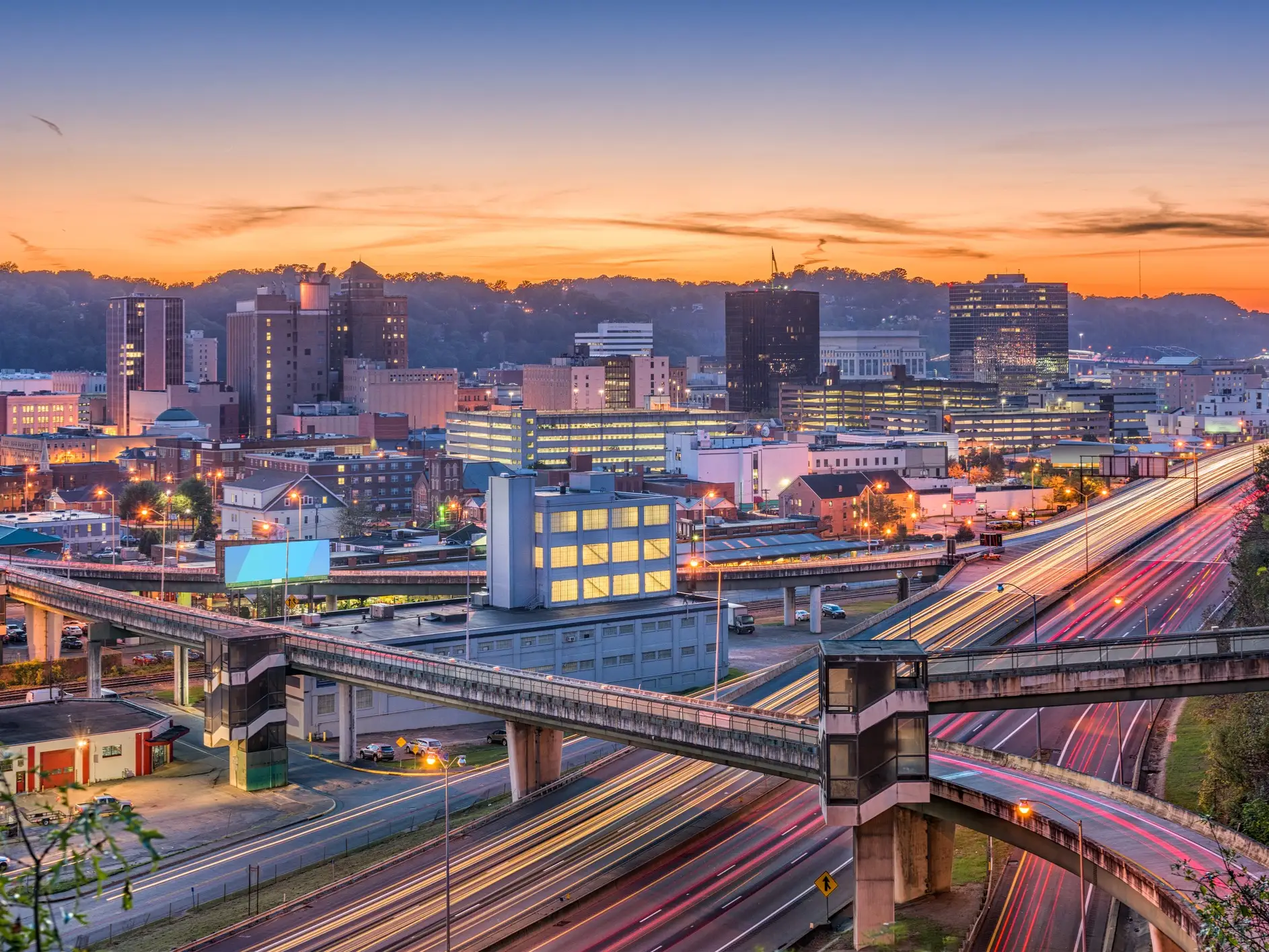 Downtown Charleston skyline at sunset with illuminated buildings, elevated walkways, highways, and long-exposure traffic trails.