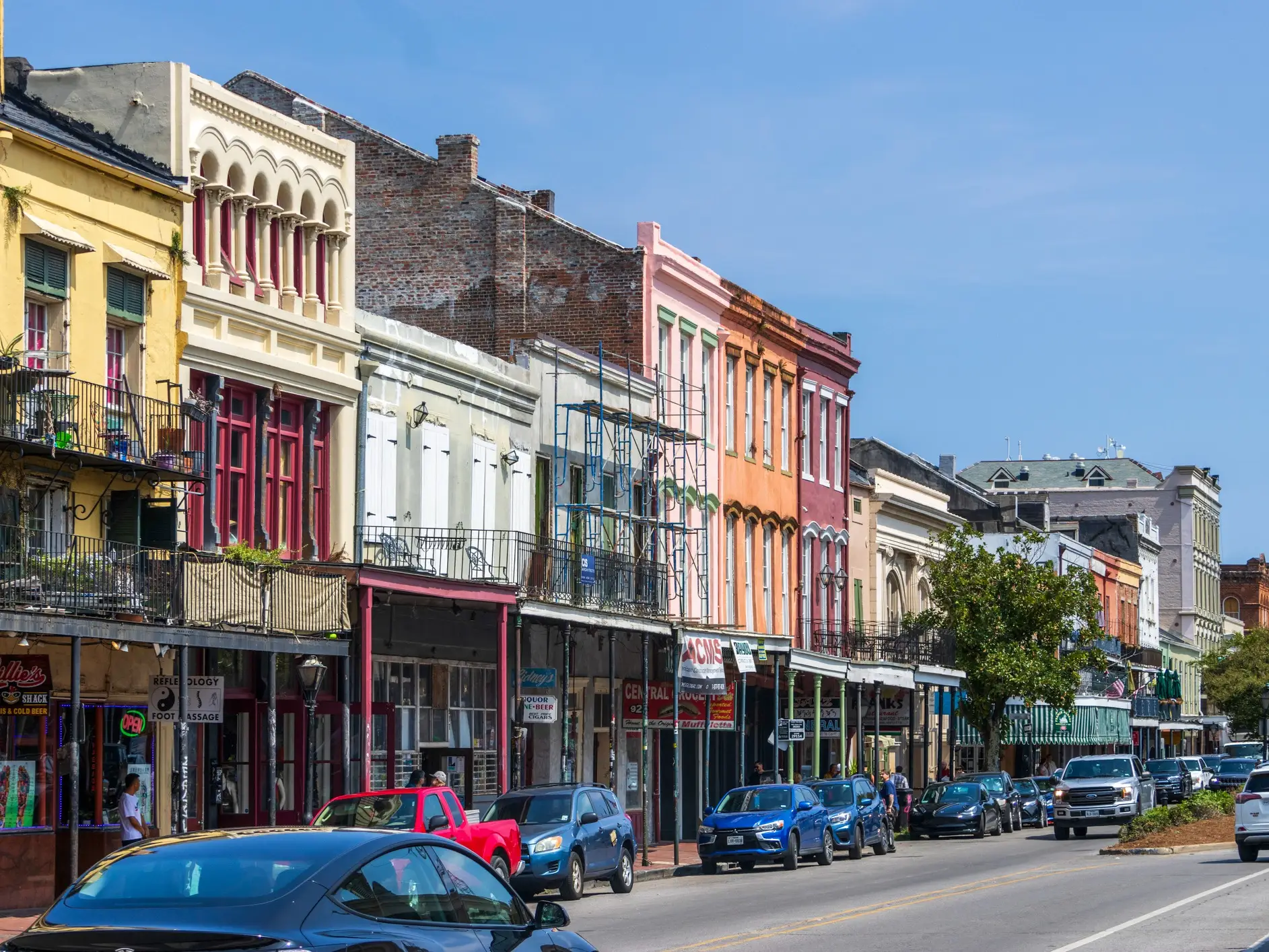 Colorful historic storefronts with balconies line a sunny city street filled with parked cars.