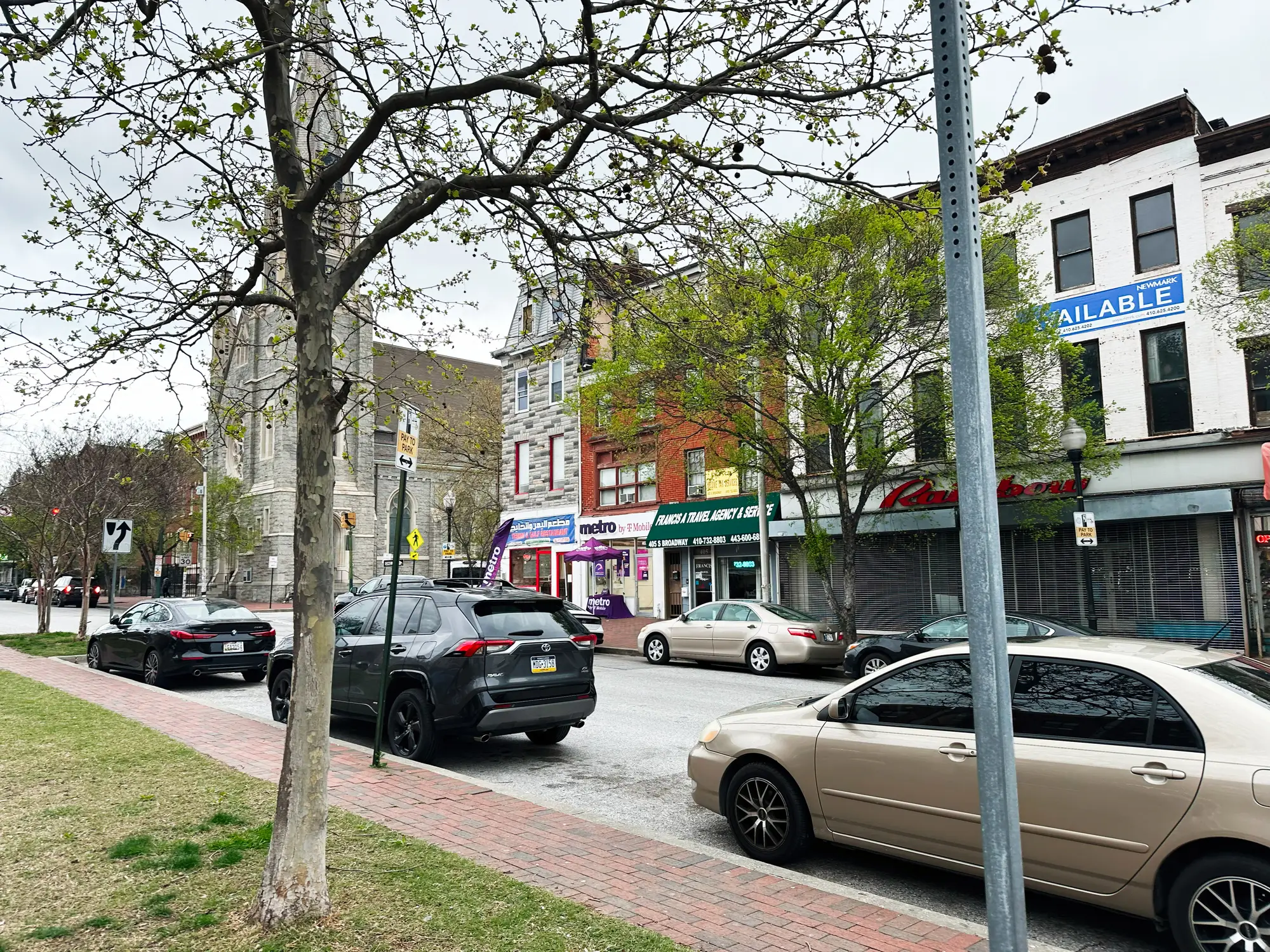Parked cars line a commercial street with storefronts, trees, and an AVAILABLE sign on an upper-floor facade.