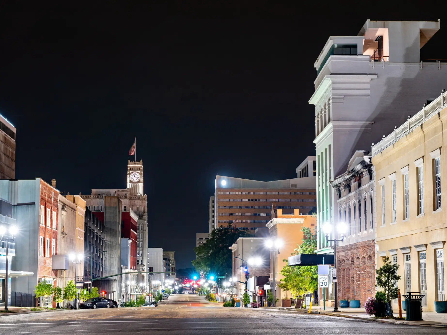Illuminated downtown street at night with historic buildings, a clock tower, streetlights, and a speed limit sign.