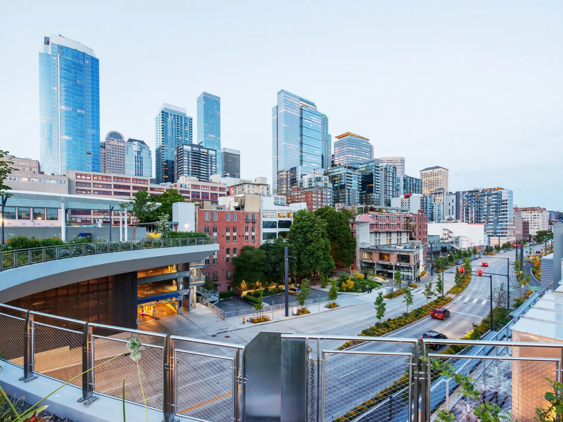 Modern glass high-rises rise above a landscaped city street and pedestrian terrace at dusk.