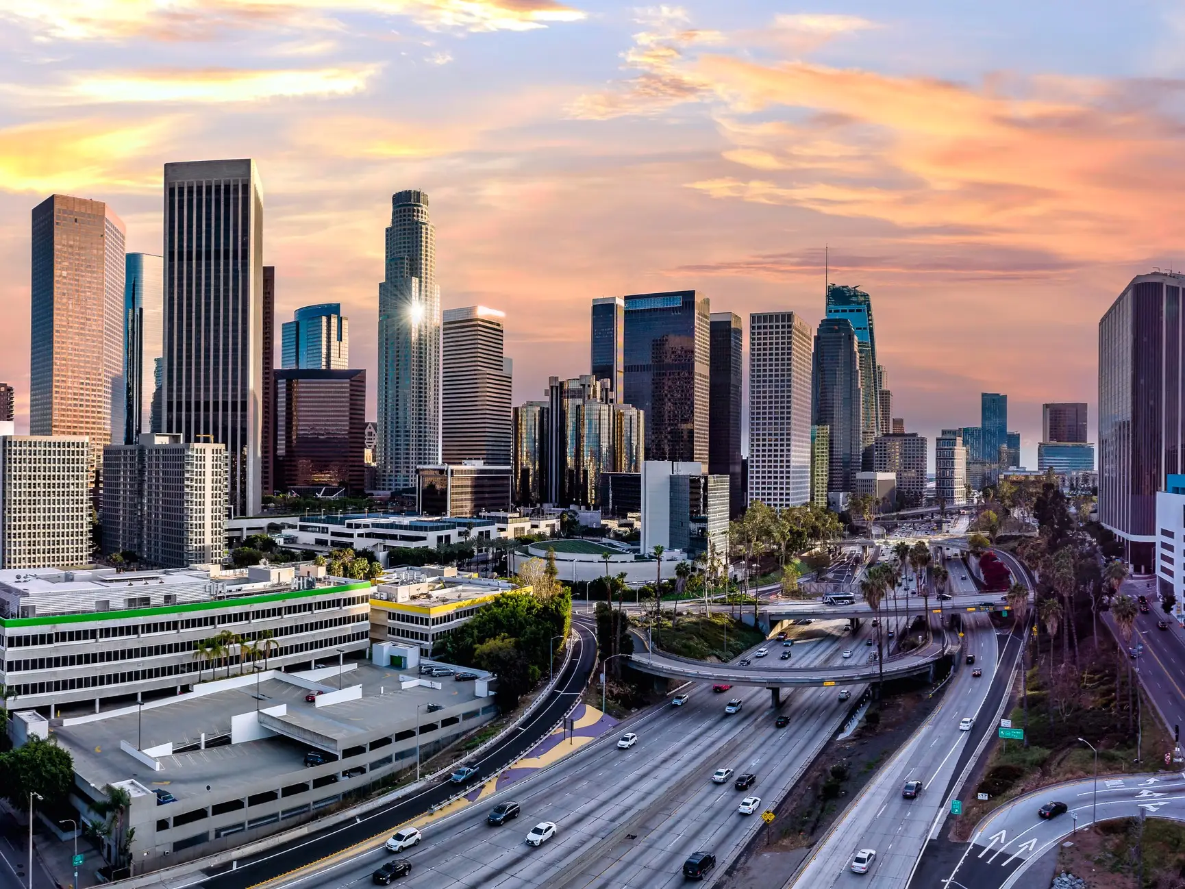Downtown Los Angeles skyline rises above multilane freeways and overpasses under a colorful sunset sky.