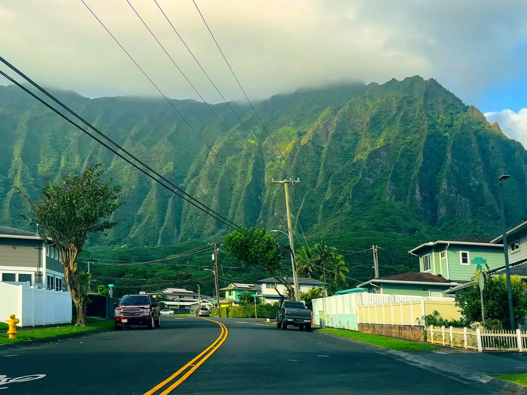 Residential street with parked vehicles and power lines beneath steep green mountains partly covered by clouds.