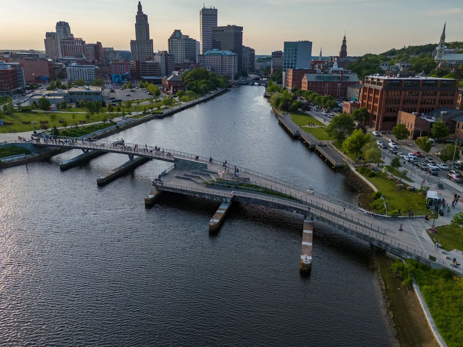Downtown Providence skyline rises beyond a pedestrian bridge over the Providence River at sunset.