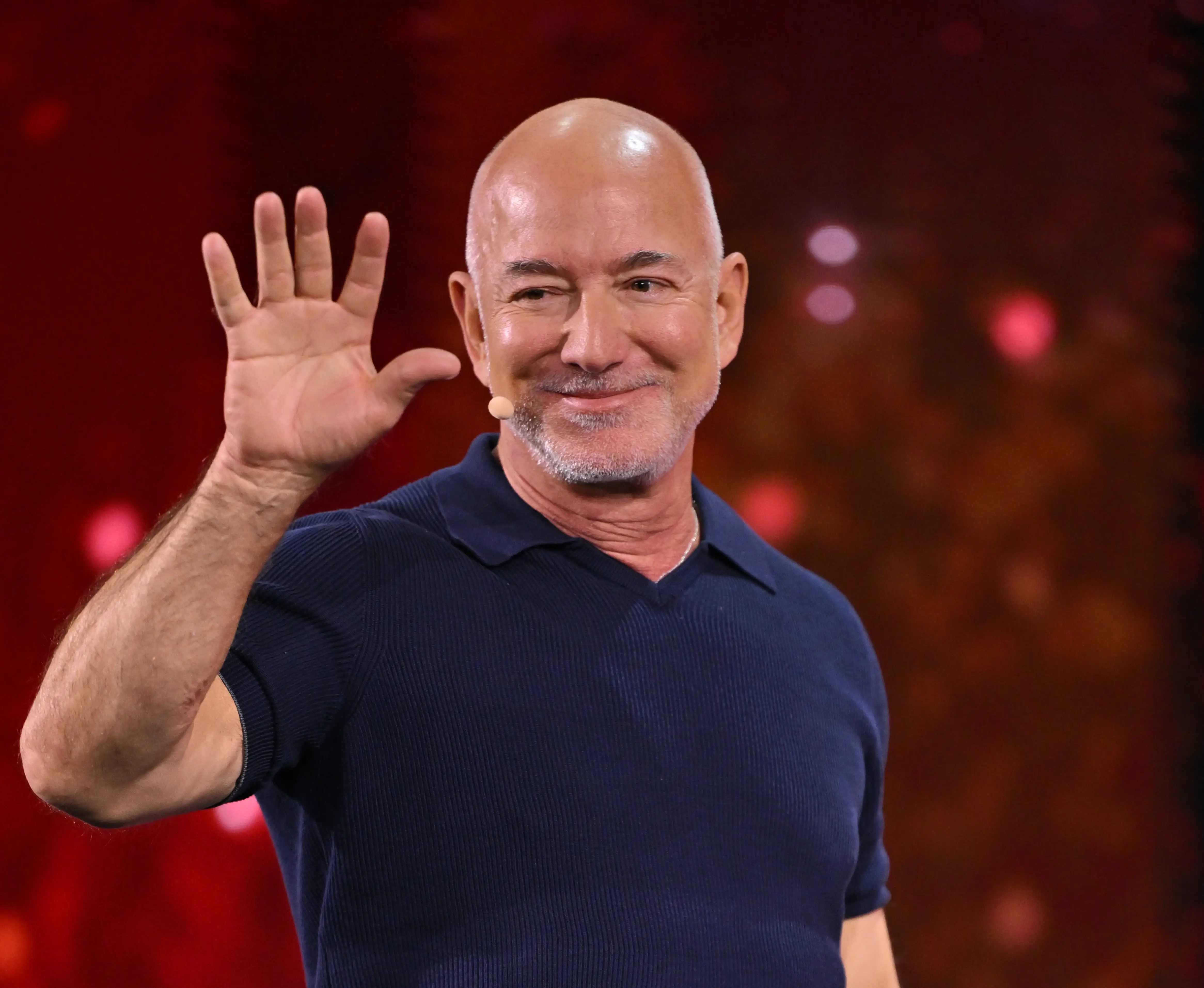 Jeff Bezos waves while standing on a stage with a red blurred background and wearing a headset microphone.