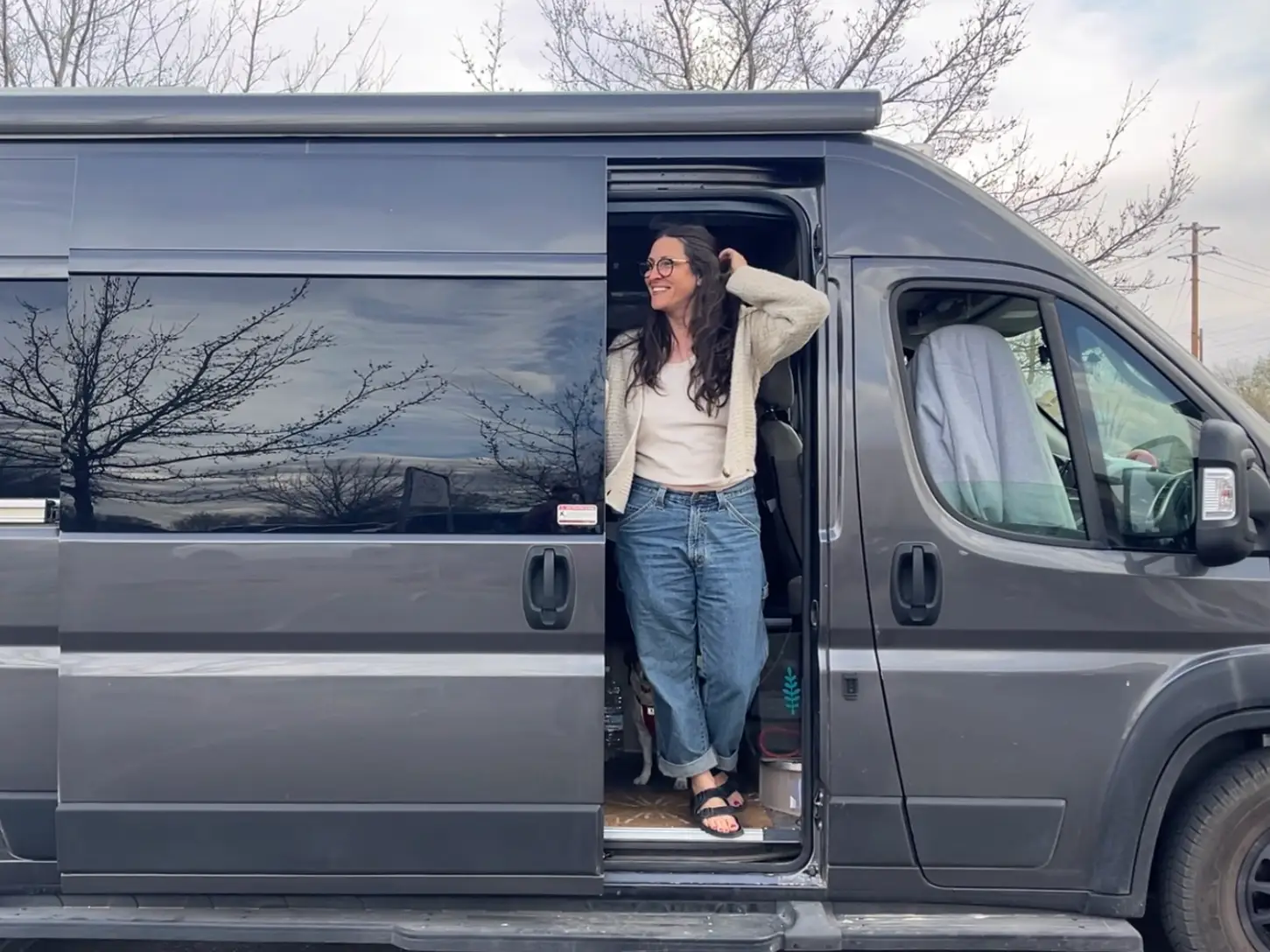 The writer stands in the open sliding doorway of a gray camper van with a small dog inside.