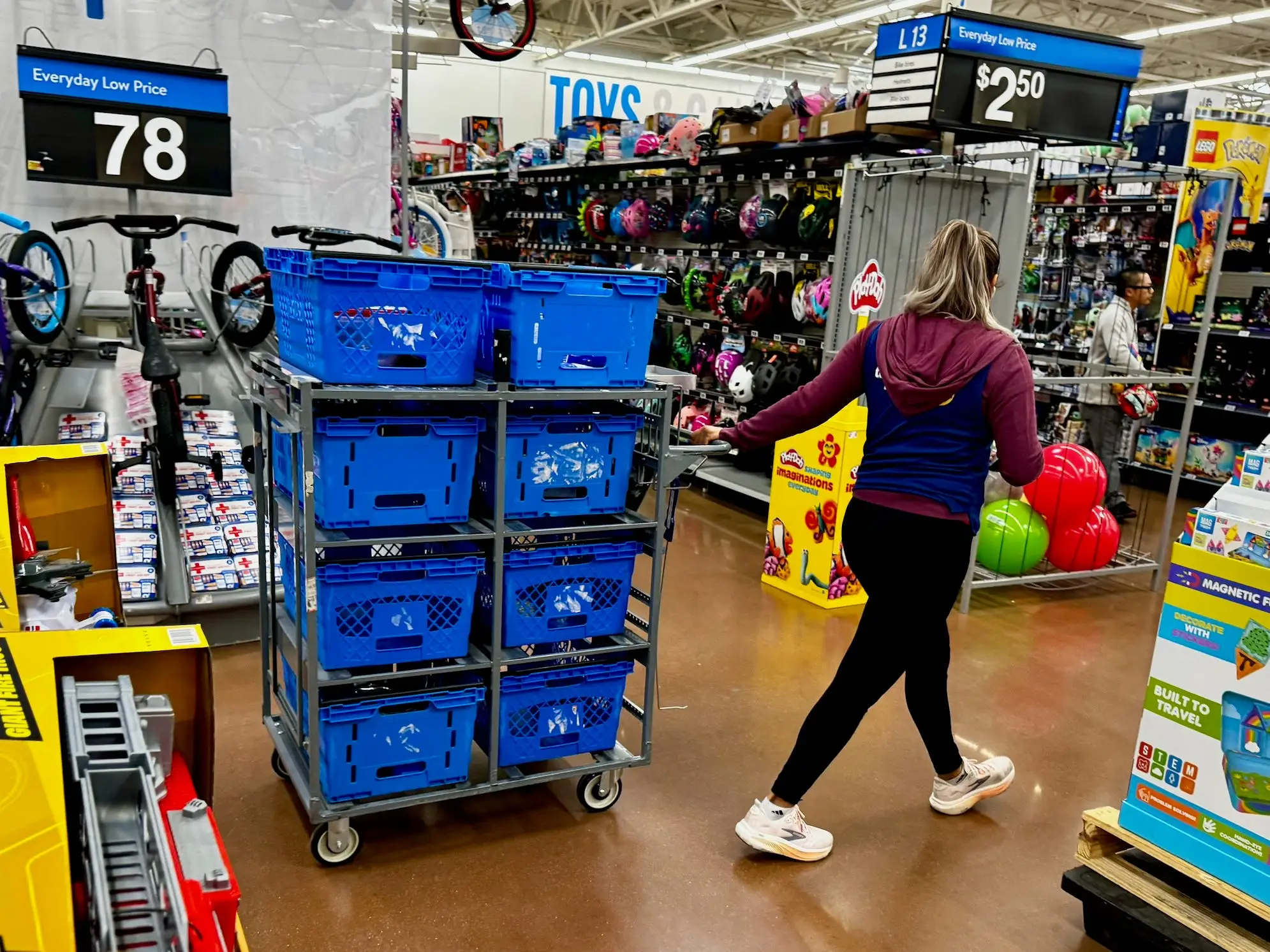 A person walks through a Walmart toy aisle with bicycles, blue bins, balls, and boxed toys on display.