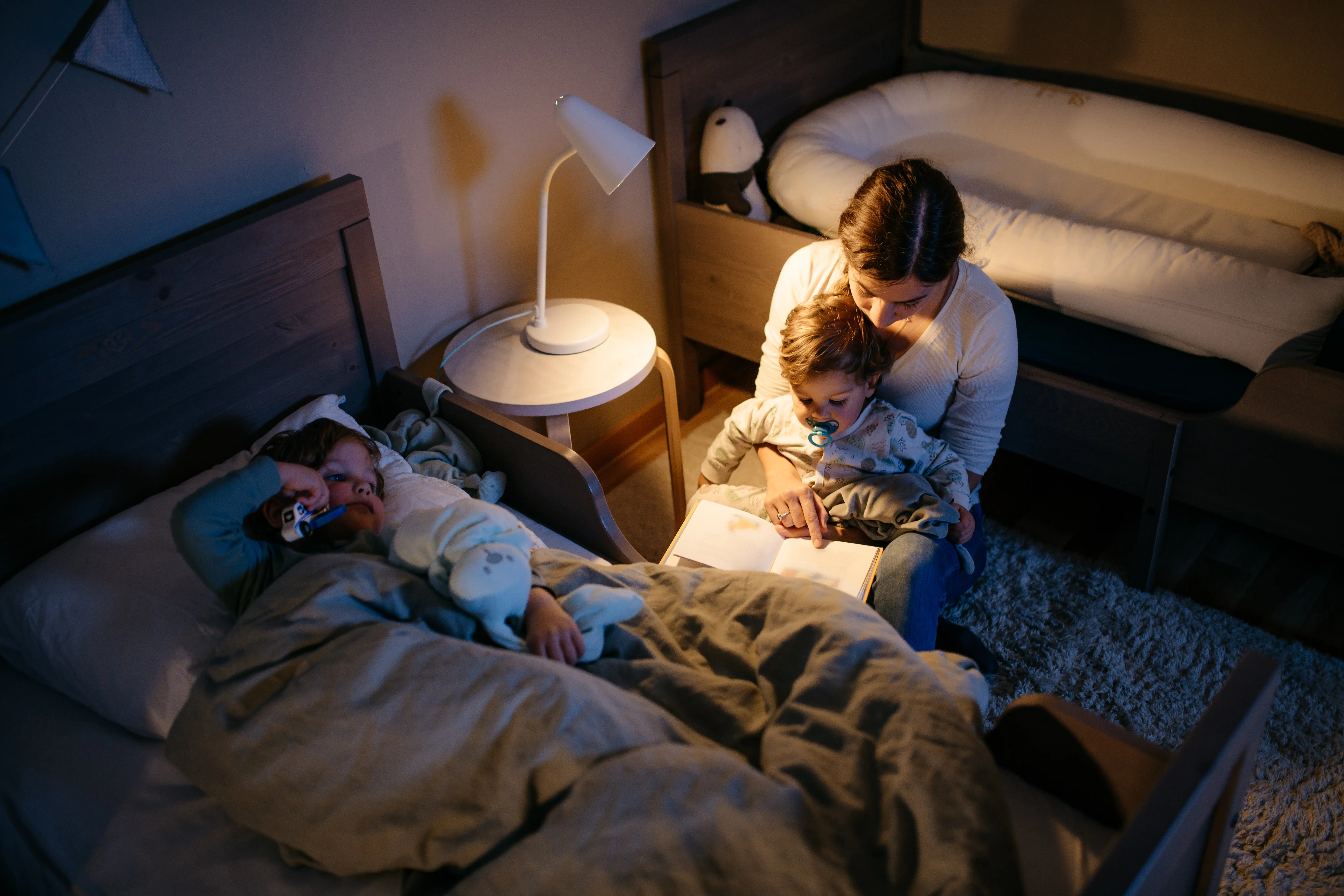 Adult reads an illuminated book beside a bed while two children rest in a dimly lit bedroom.