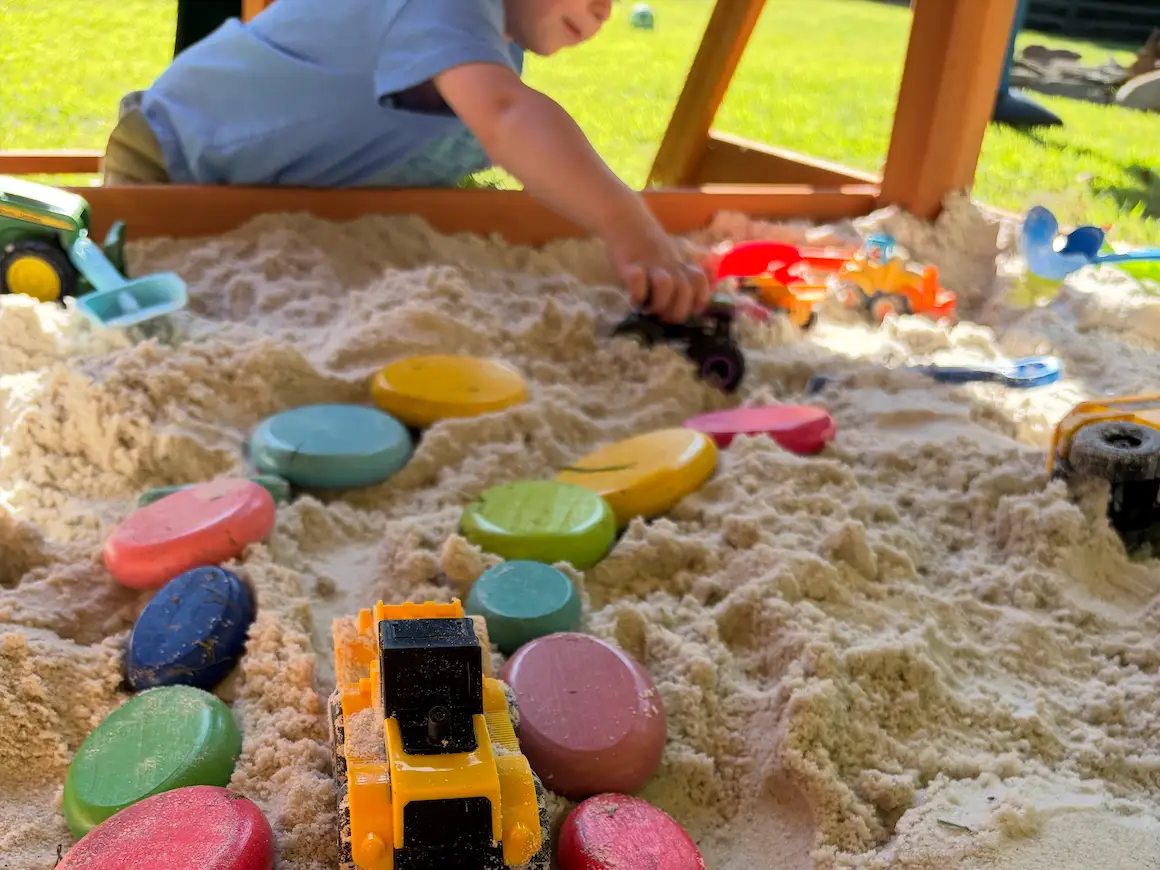 A person reaches across a sandbox filled with colorful toy stones, vehicles, and sand tools outdoors.