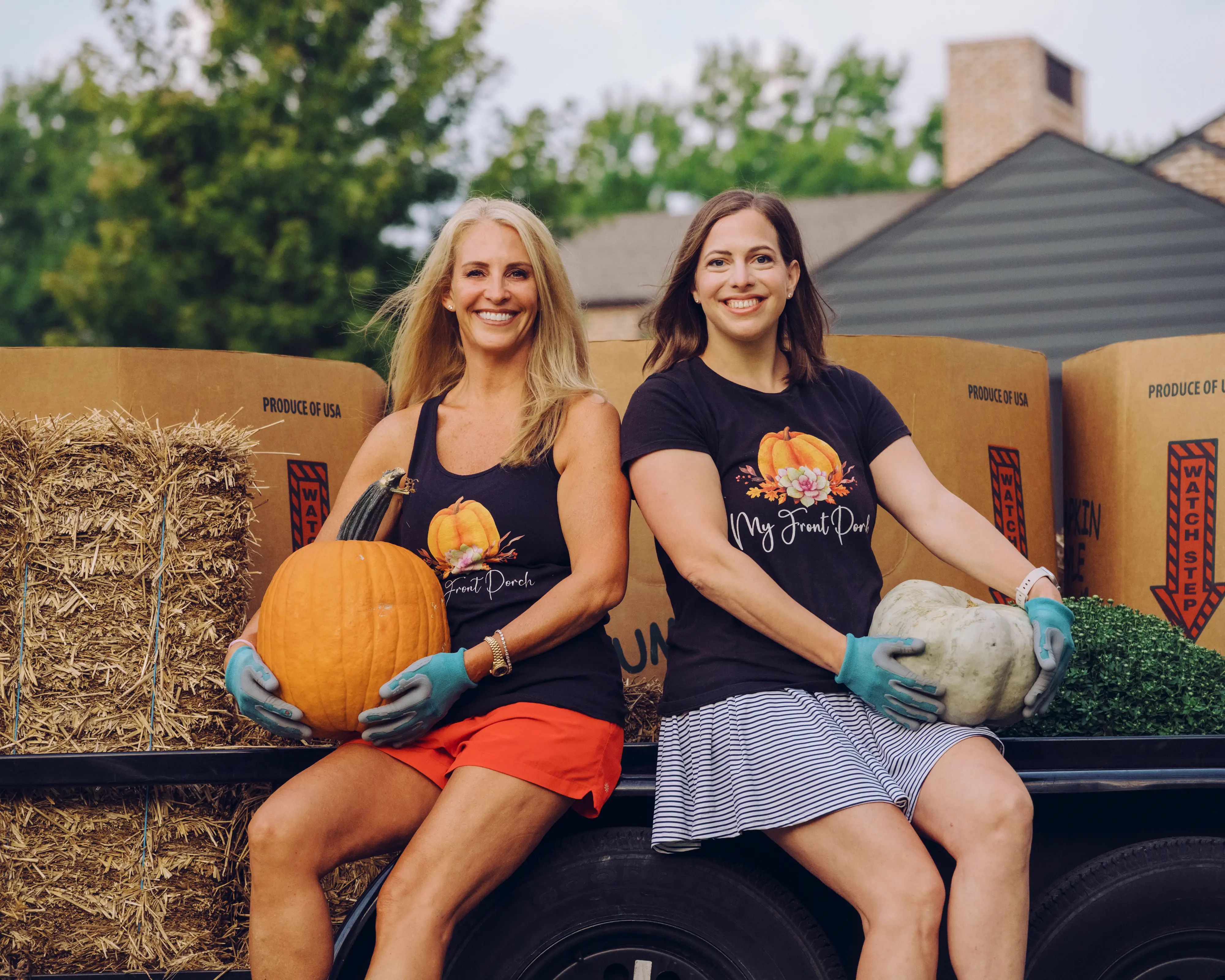Jen and Rebecca sit on a trailer holding pumpkins among hay bales and produce boxes outdoors.