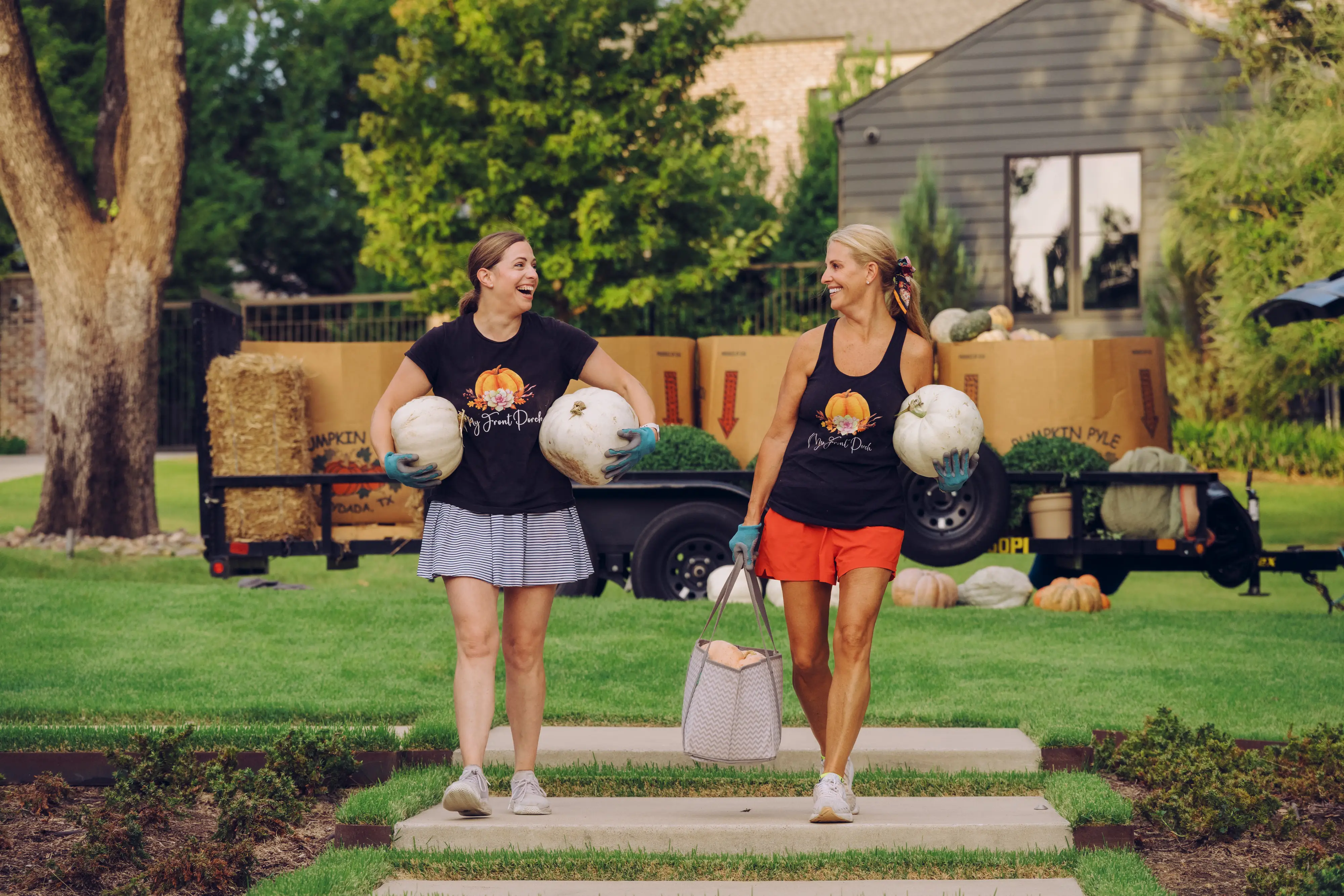 Rebecca and Jen carry white pumpkins past trailers of fall decor on a residential lawn.