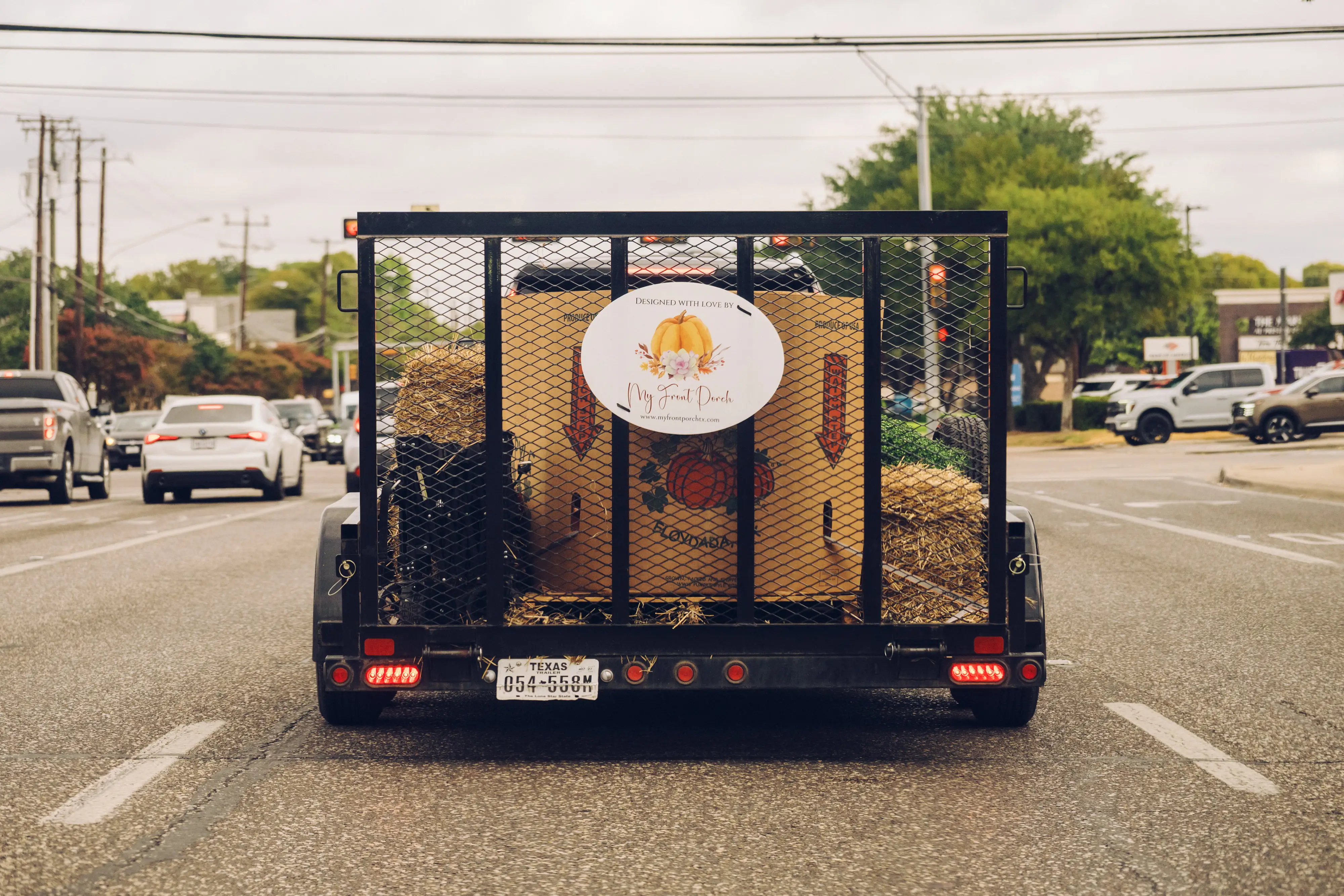 A trailer carrying hay bales and pumpkin decorations drives along a multi-lane road in traffic.