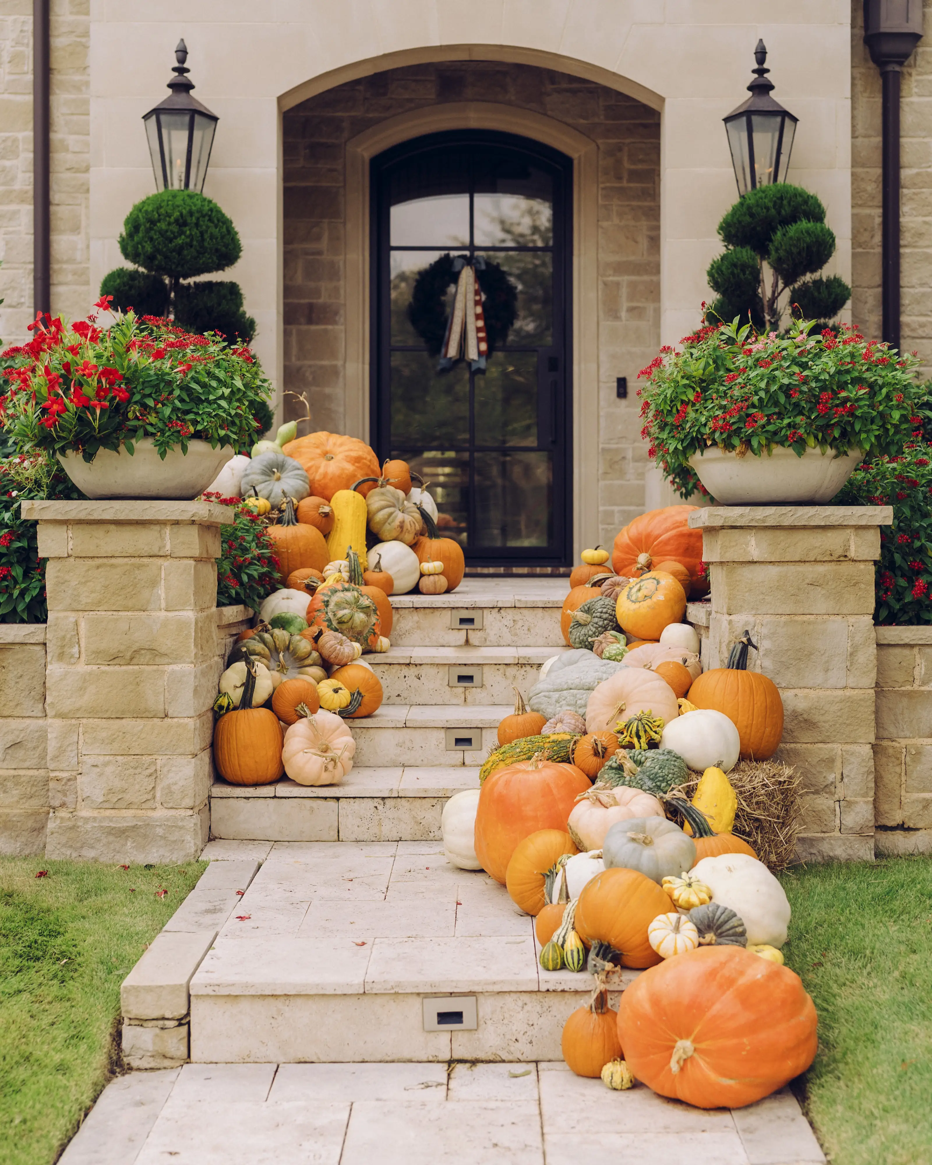 Stone house entry steps are decorated with pumpkins, gourds, red flowers, topiary planters, and an autumn wreath.