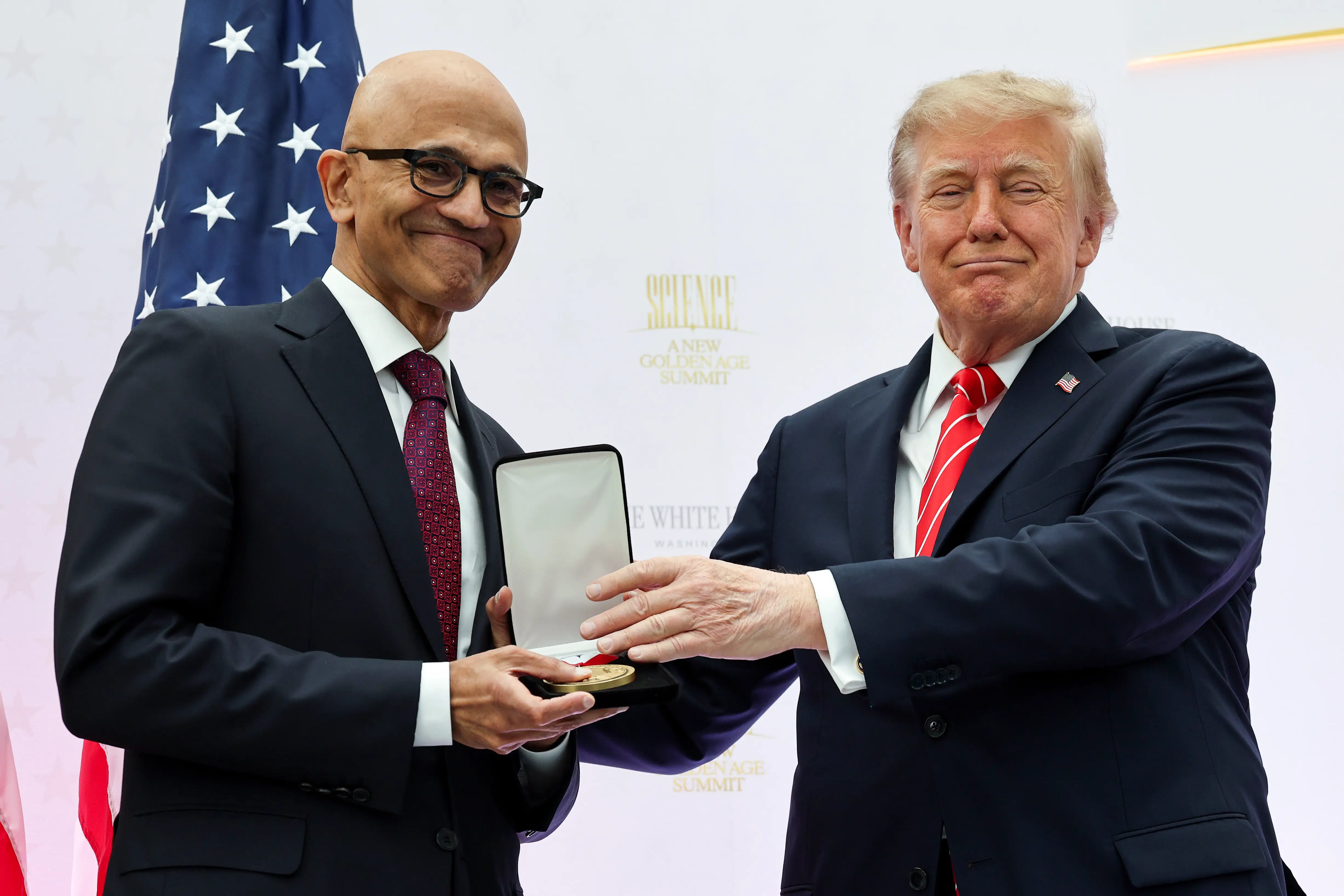 Satya Nadella holds an open medal case while Donald Trump gestures beside him with a U.S. flag behind them.
