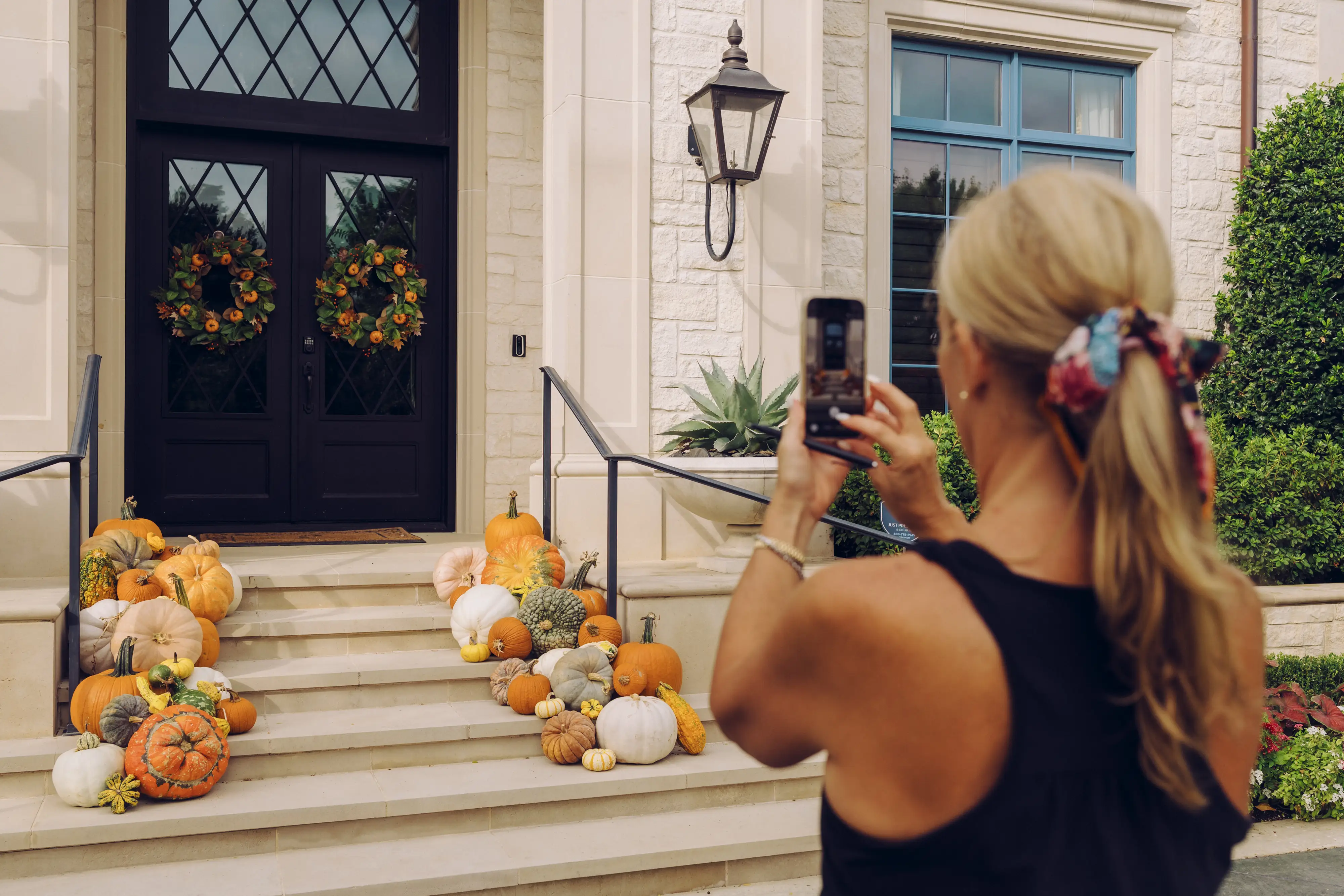 Jen photographs a home's front steps decorated with pumpkins, gourds, and fall wreaths using a smartphone.