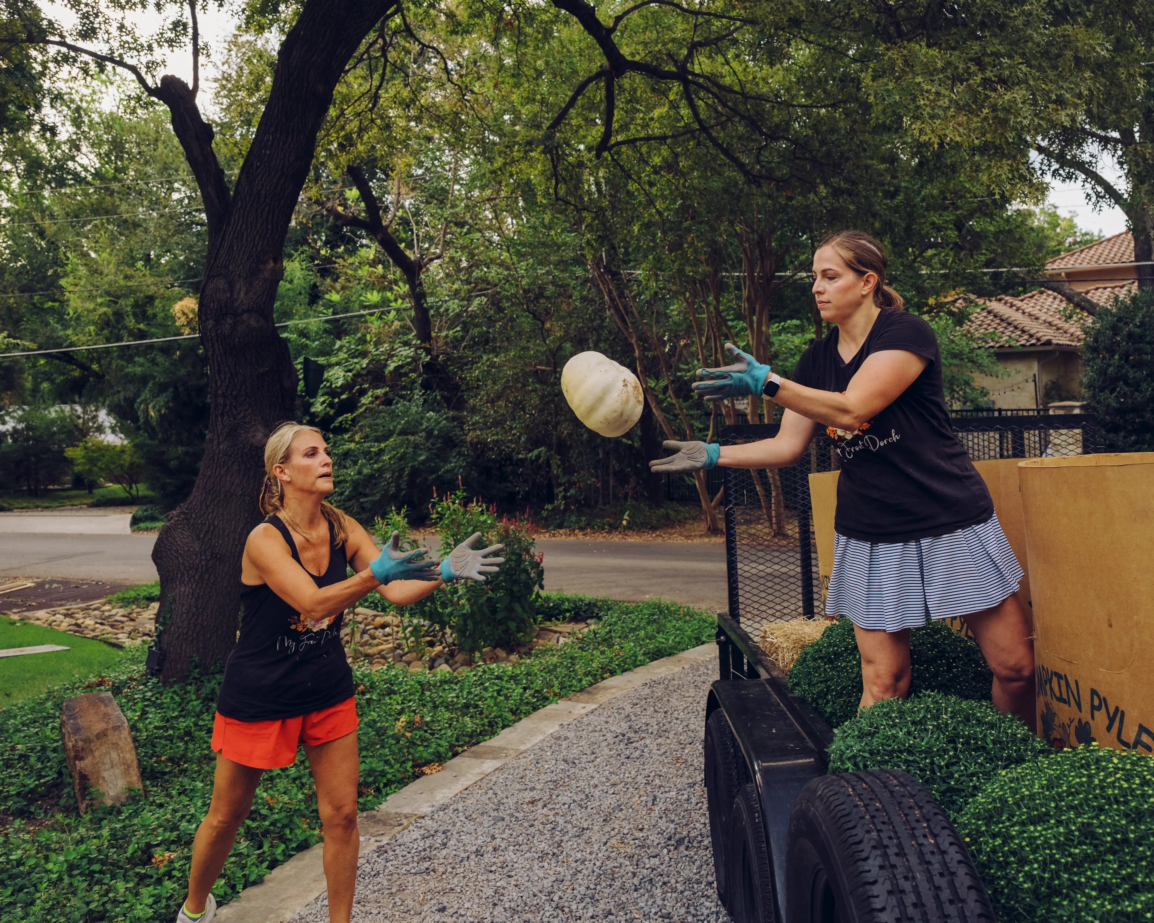 Rebecca and Jen wearing gloves toss a pale pumpkin beside a landscaping trailer in a leafy residential yard.
