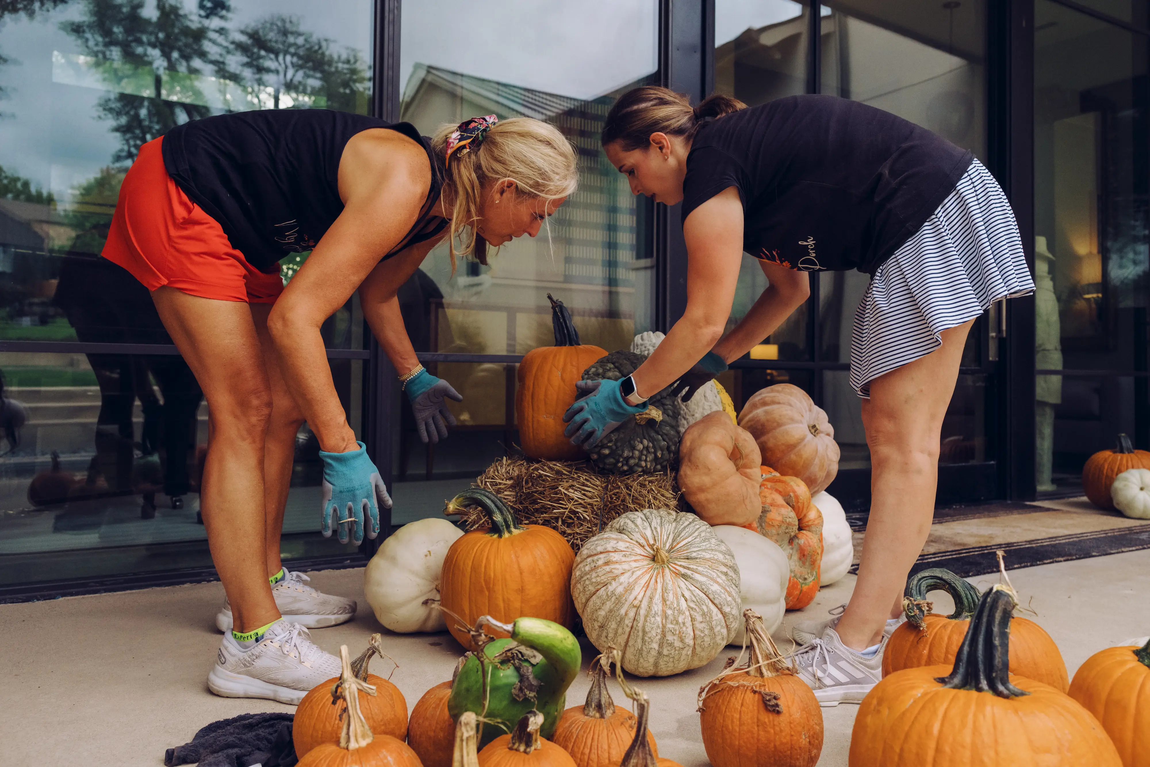 Jen and Rebecca wearing gloves arrange pumpkins and gourds on a porch beside large glass doors.
