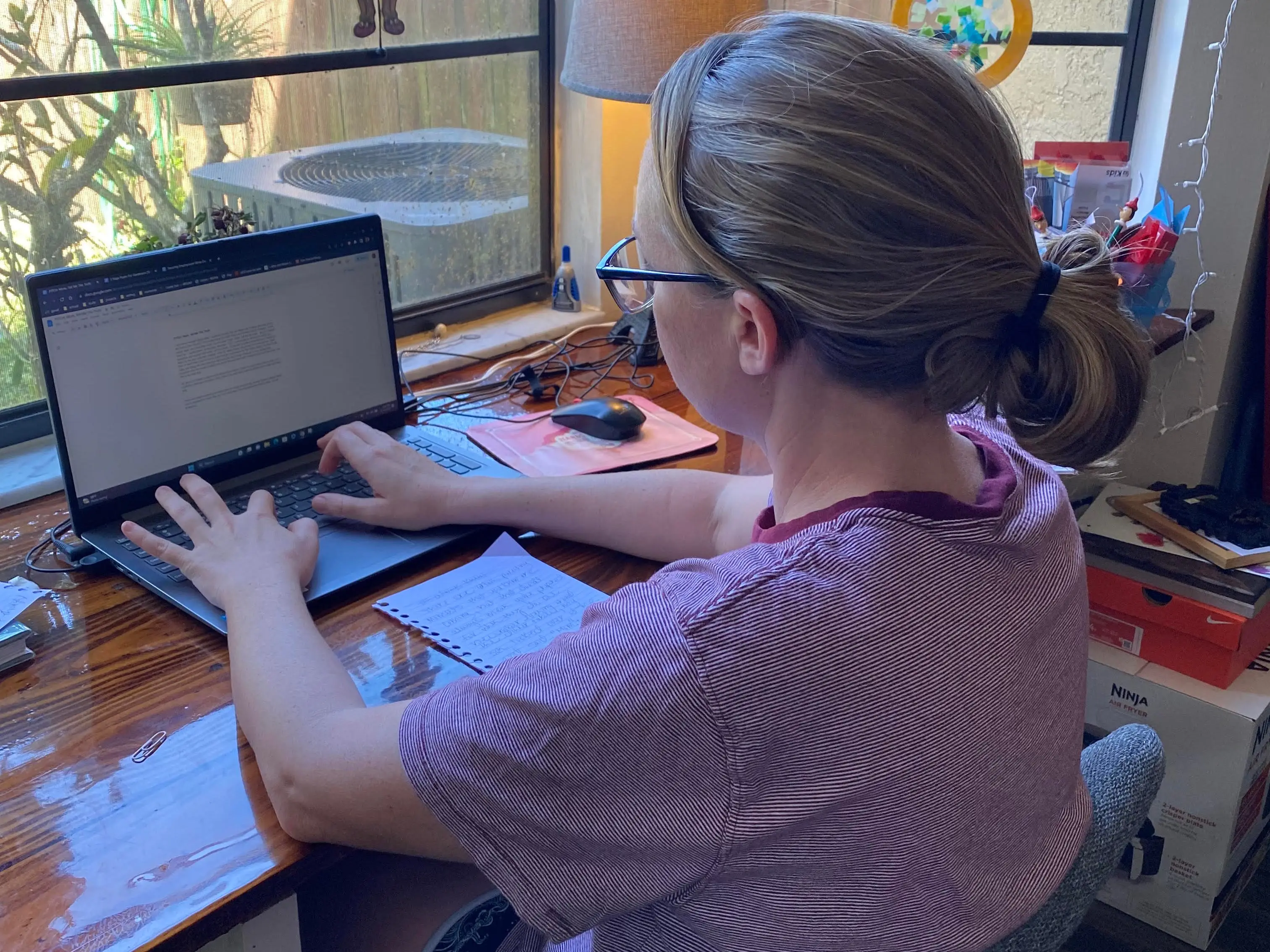 Person wearing glasses works on a laptop at a wooden desk beside large sunlit windows.