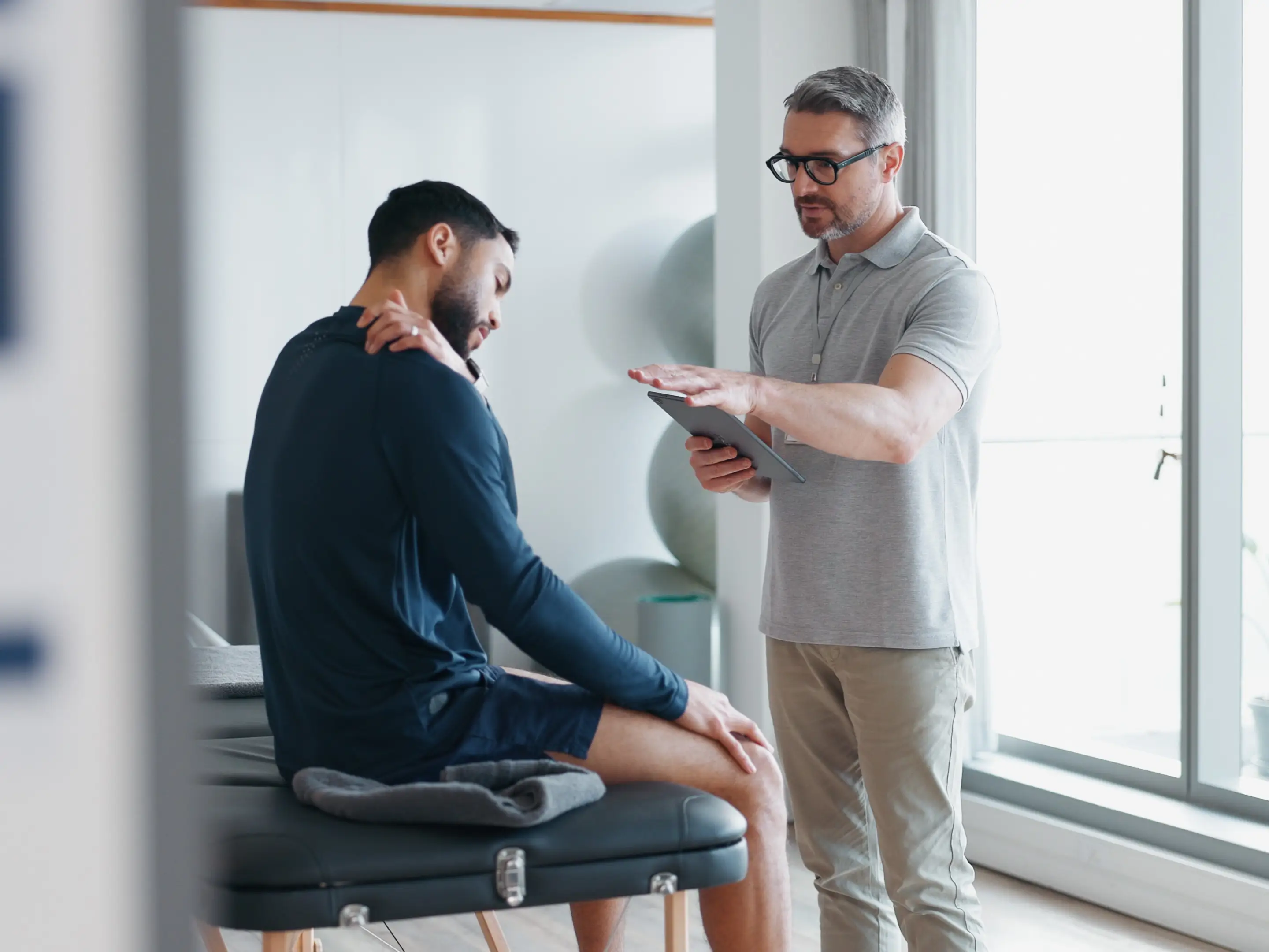 Physiotherapy, tablet and shoulder pain with people in clinic for rehabilitation, healthcare or consulting. Support, medical and chiropractor with man and athlete for healing, advice or report stock photo