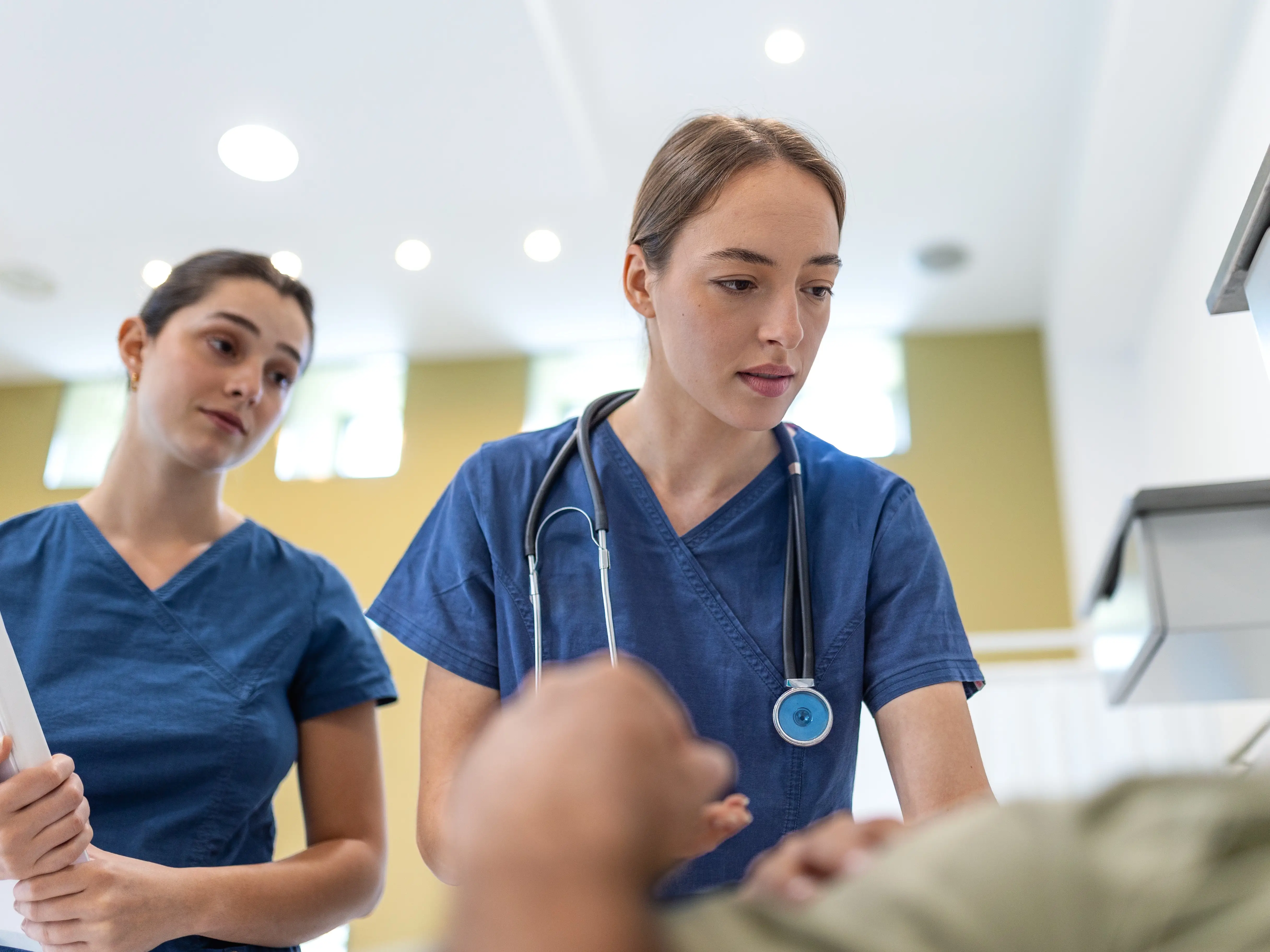 Two focused female doctors in blue scrubs checking on a senior male patient lying in a hospital bed, with one practitioner leaning forward with a stethoscope while the other holds medical charts. stock photo