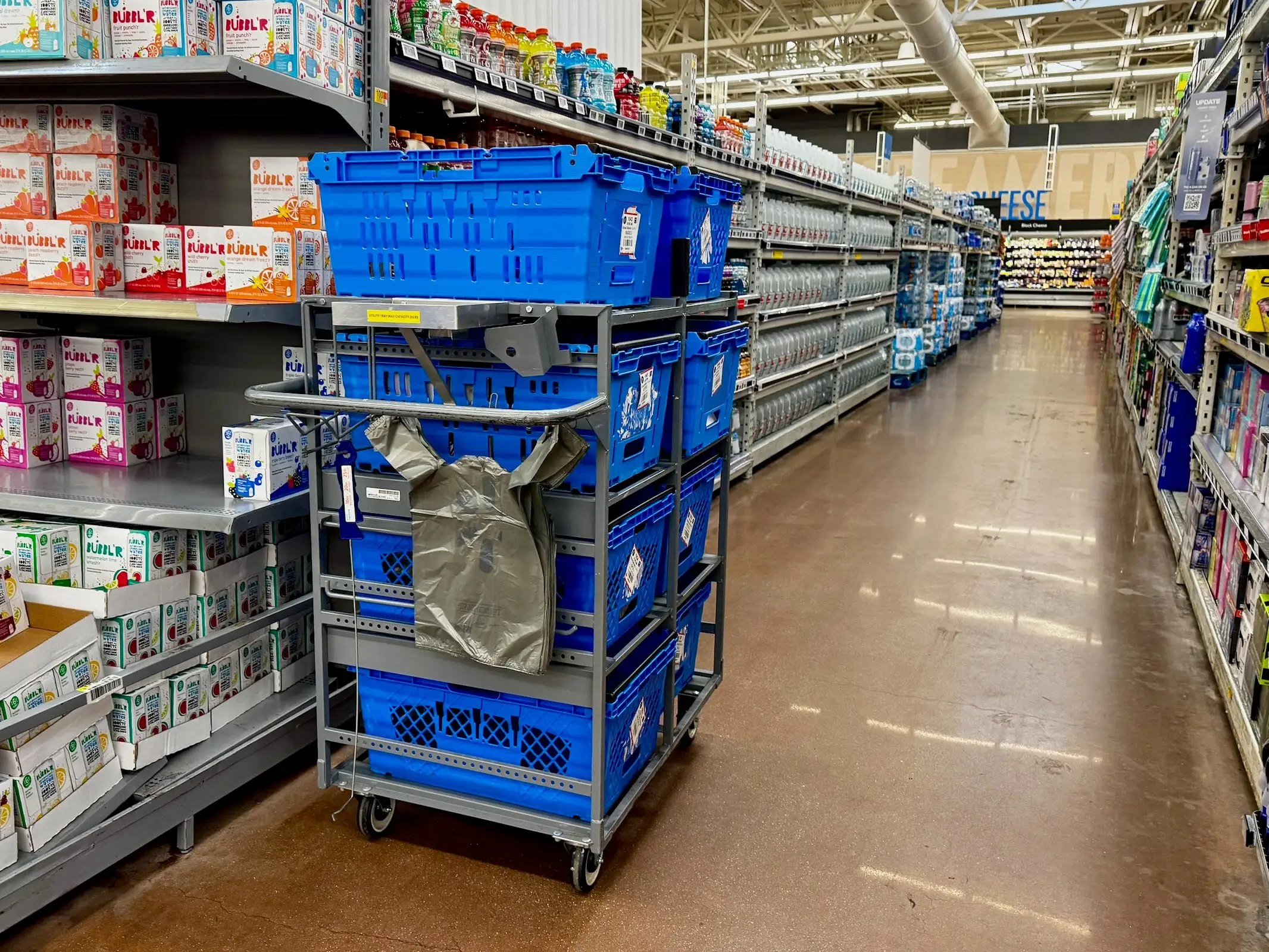 Grocery store aisle with beverage cartons on shelves and blue crates stacked on a rolling cart.