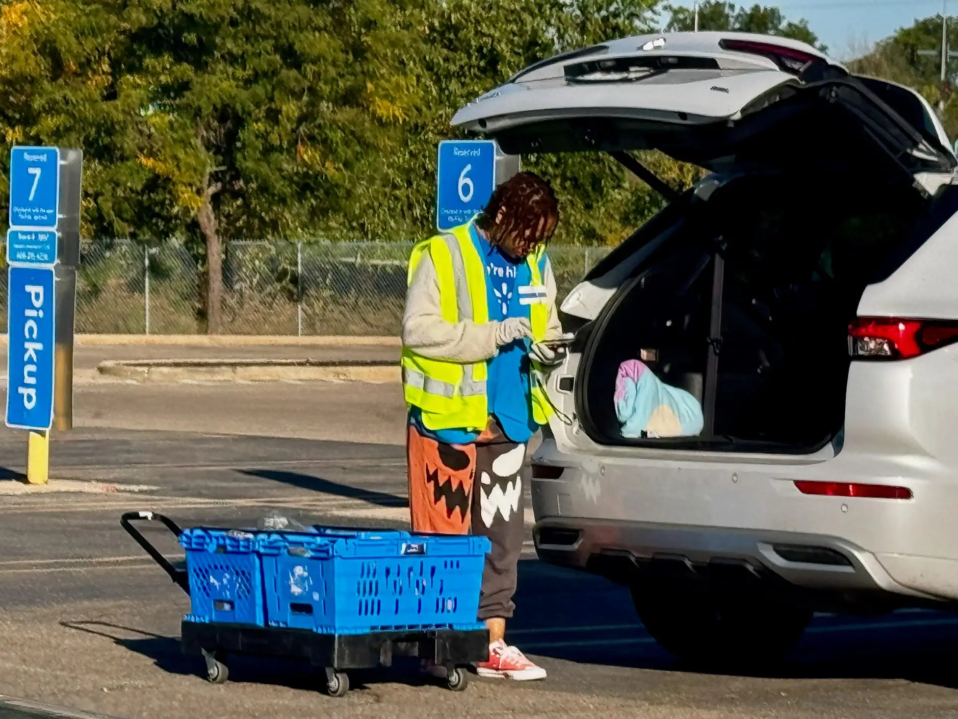 Walmart pickup worker stands beside an open SUV trunk with blue bins in a curbside pickup area.