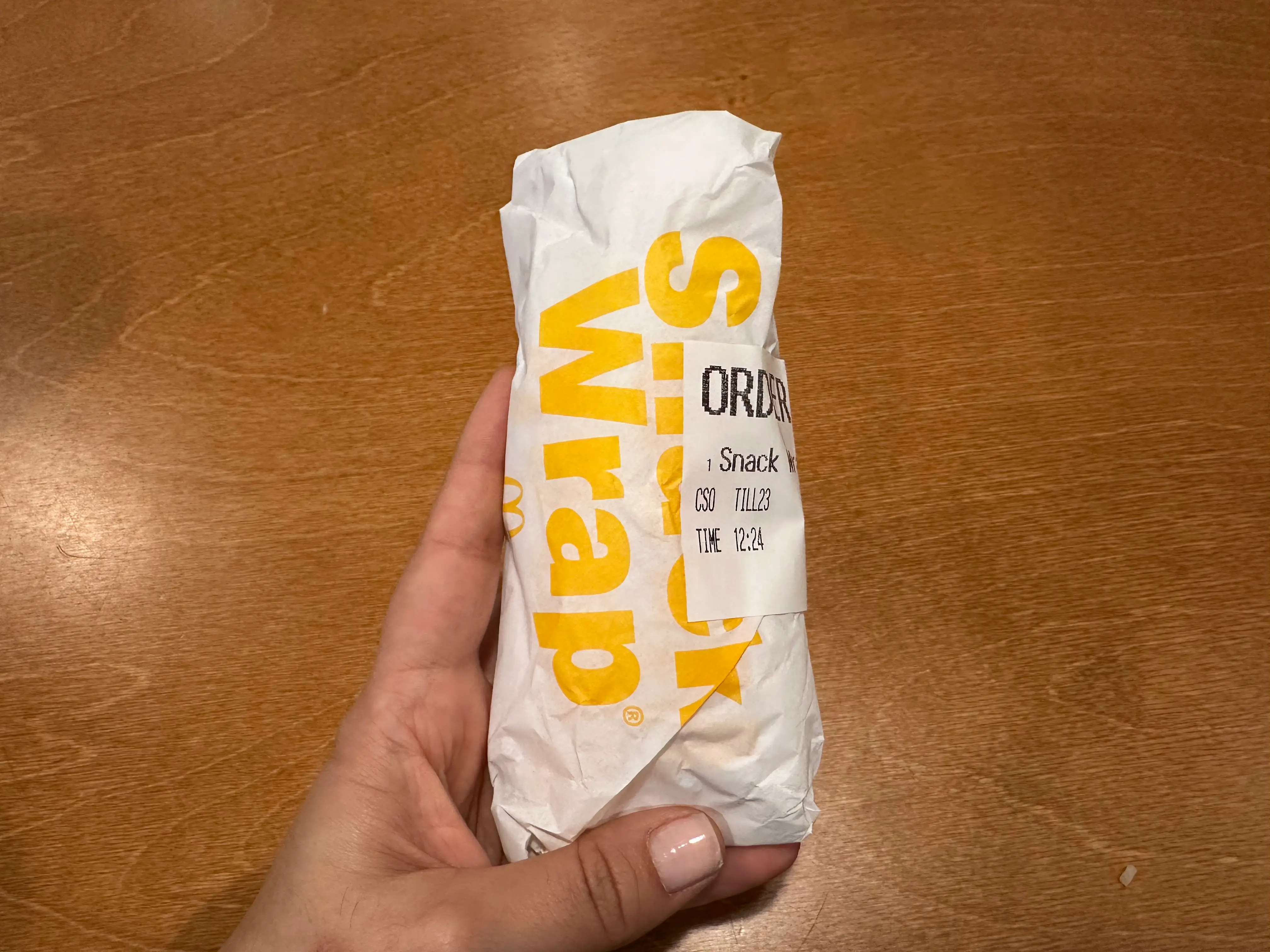 Hand holding a Subway wrapped food item with an order label on a wooden table.