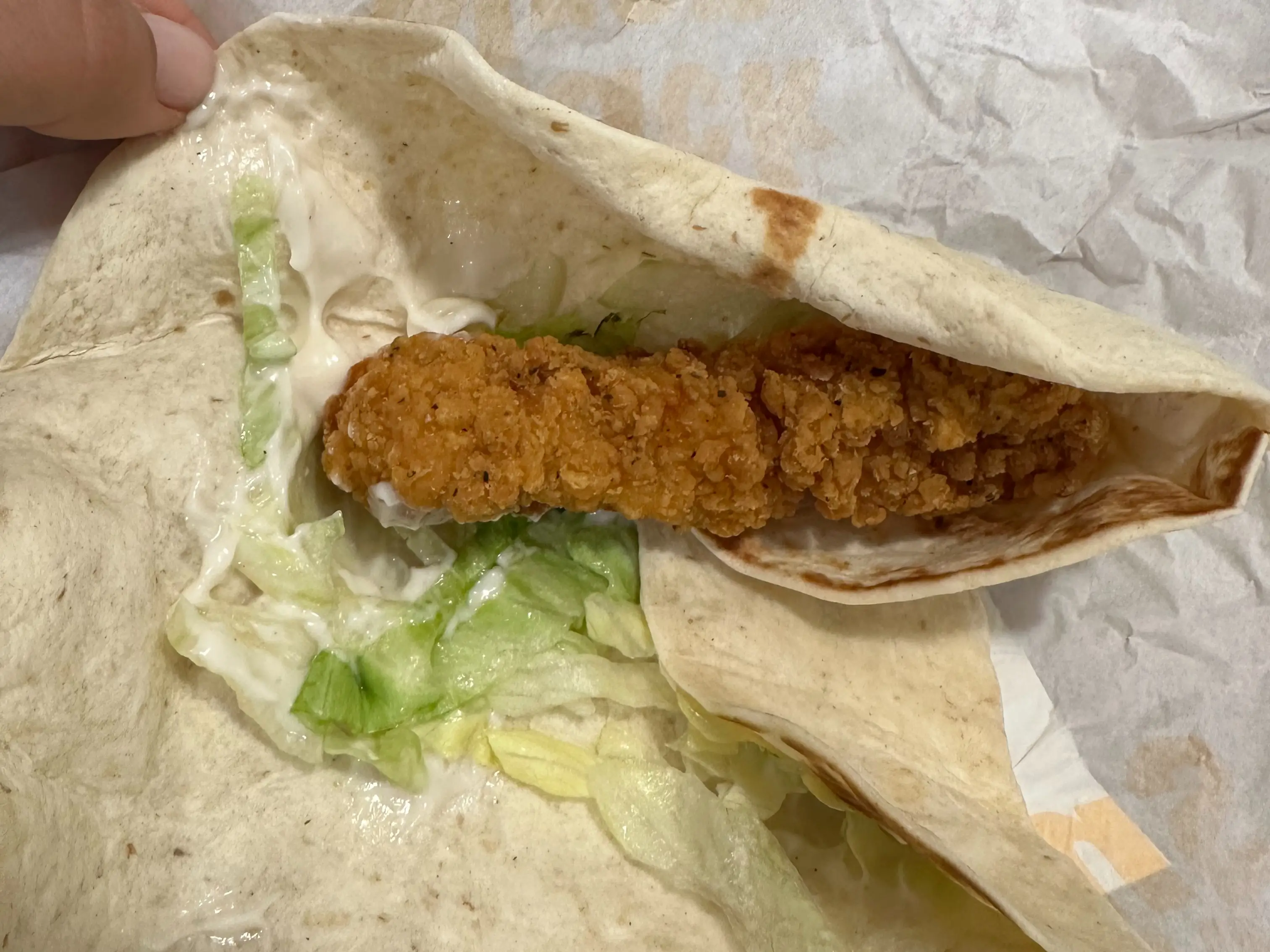 A crispy chicken strip wrap with lettuce and sauce is unfolded on fast-food wrapping paper.