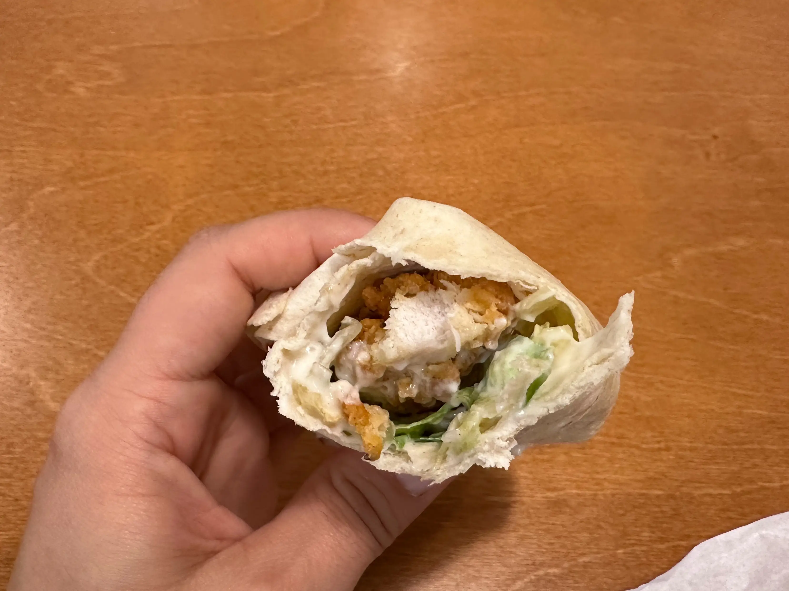 Hand holding a partially eaten tortilla wrap with lettuce and sauce over a wooden table.