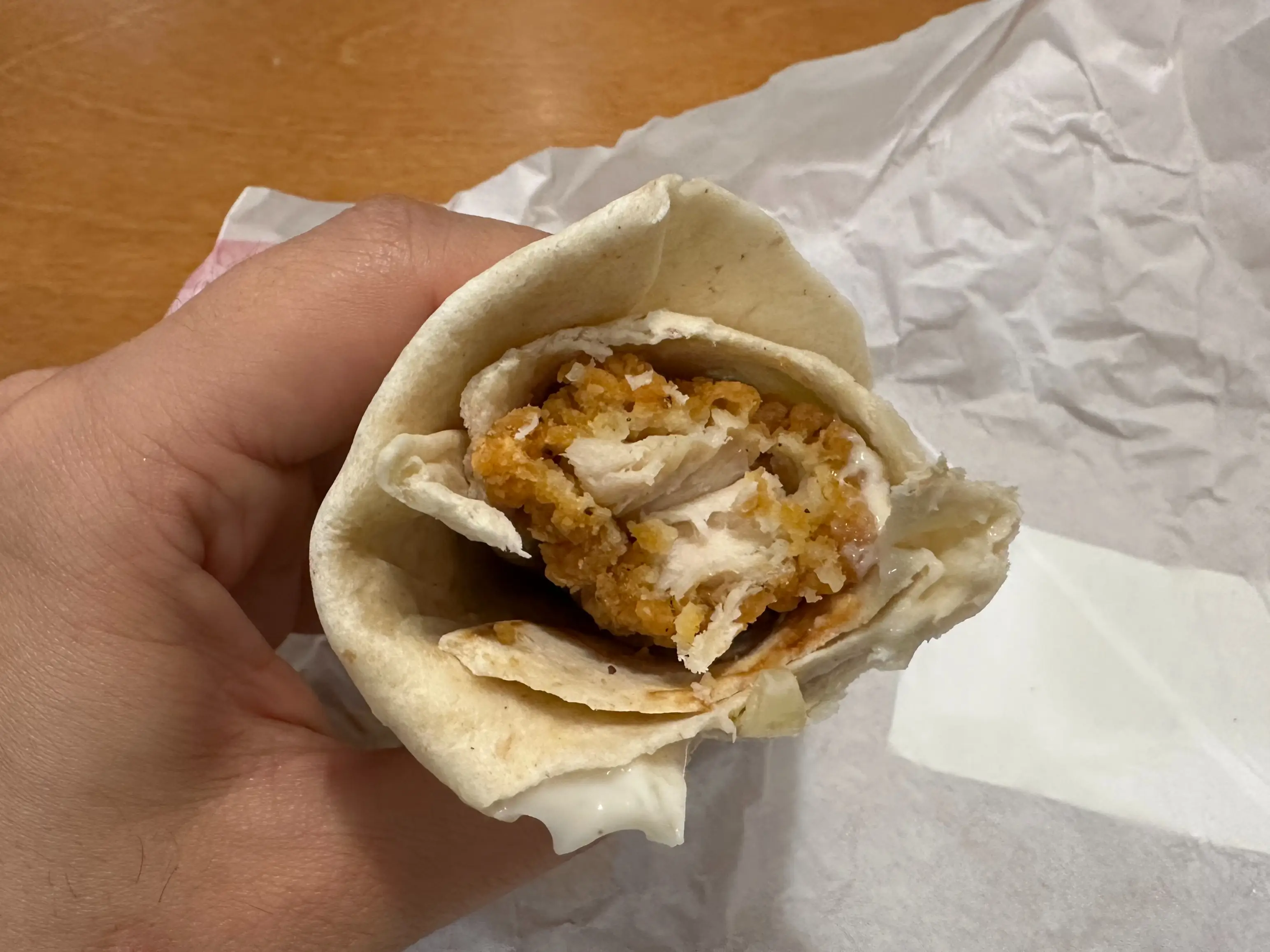 A hand holds a bitten fried chicken wrap over crumpled white paper on a wooden table.