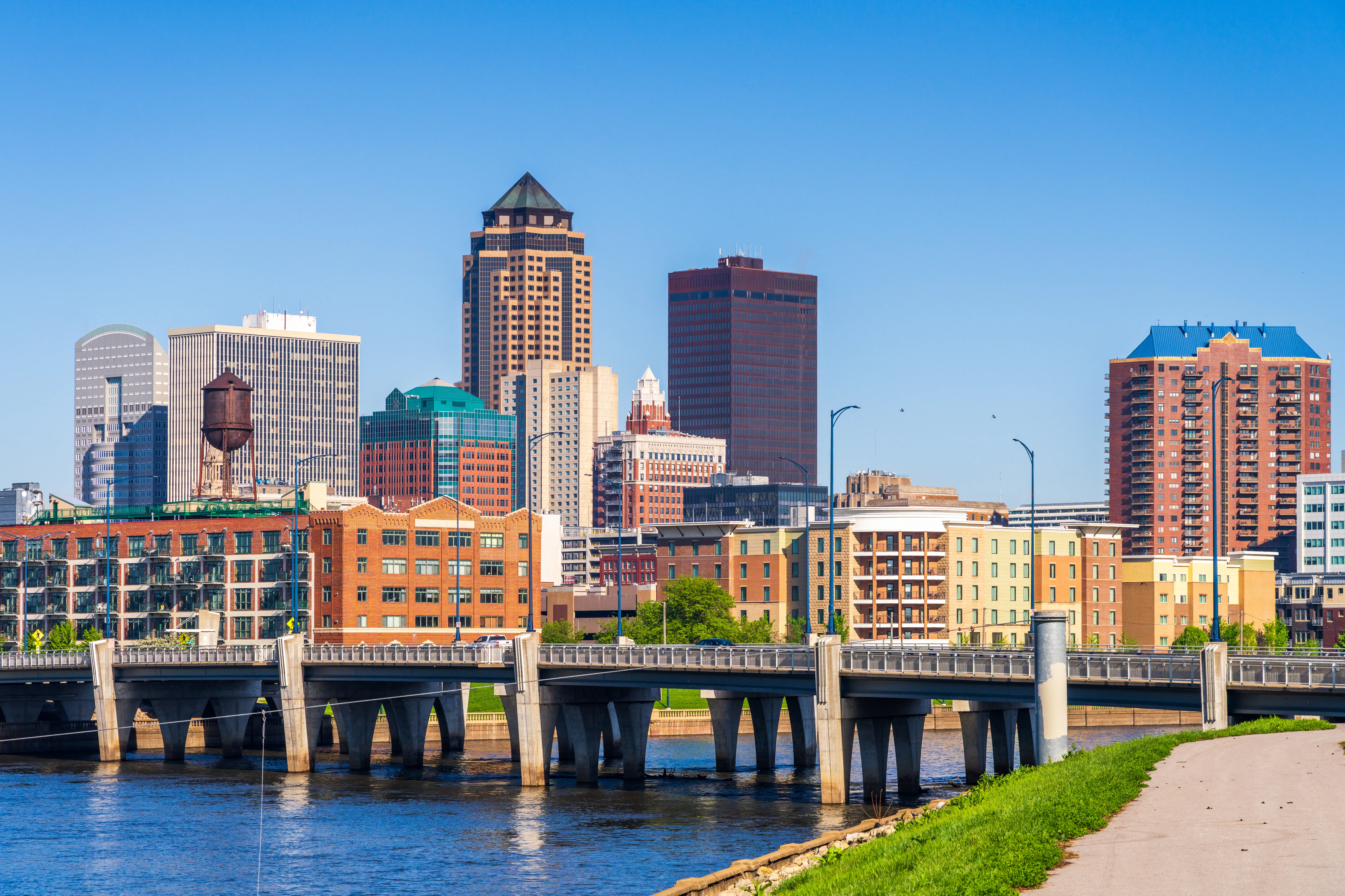 Downtown Des Moines skyline rises beyond a riverfront bridge and walkway under a clear blue sky.