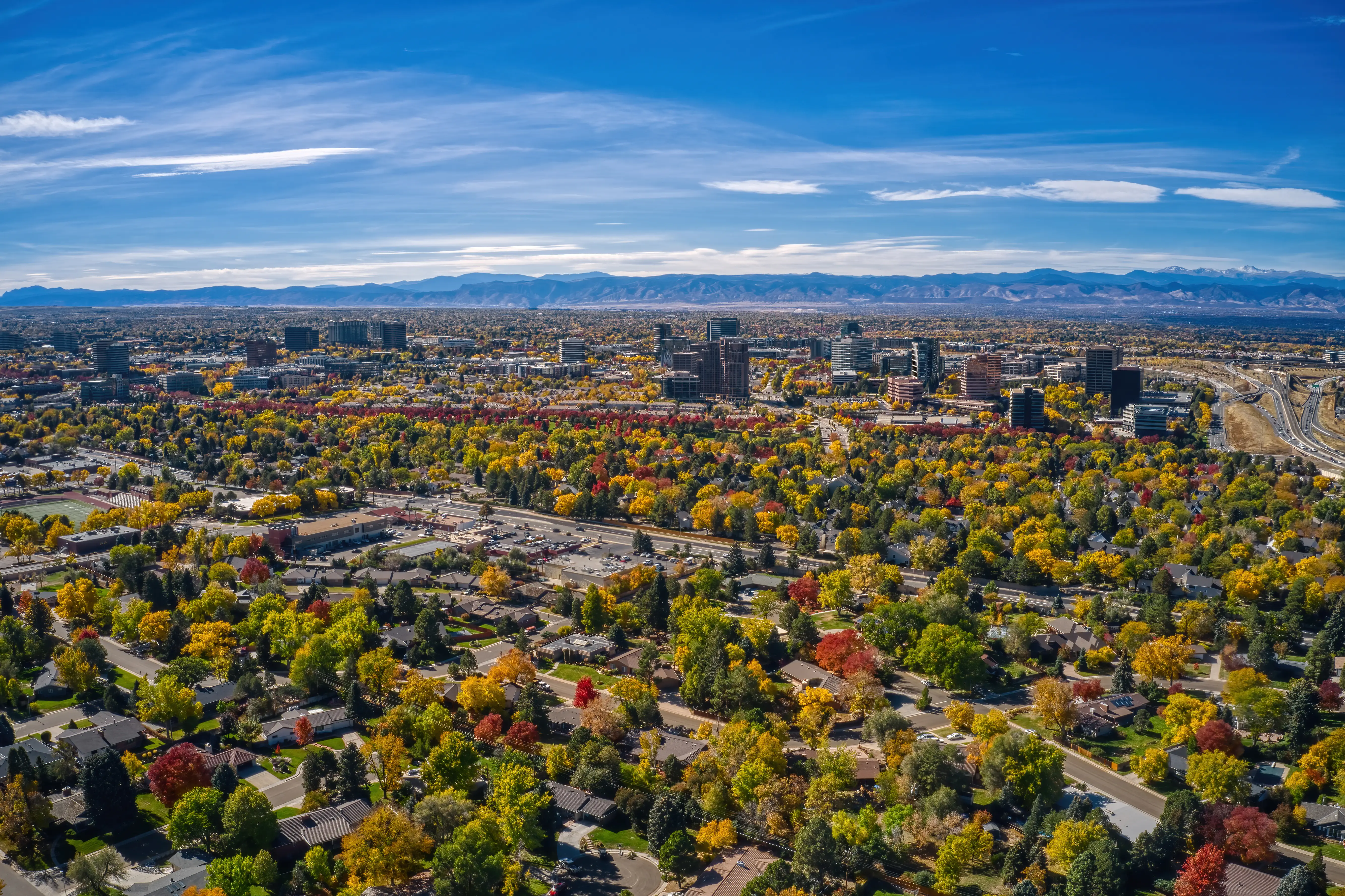 Aerial view of a suburban cityscape with autumn trees, mid-rise buildings, highways, and distant mountains under a blue sky.
