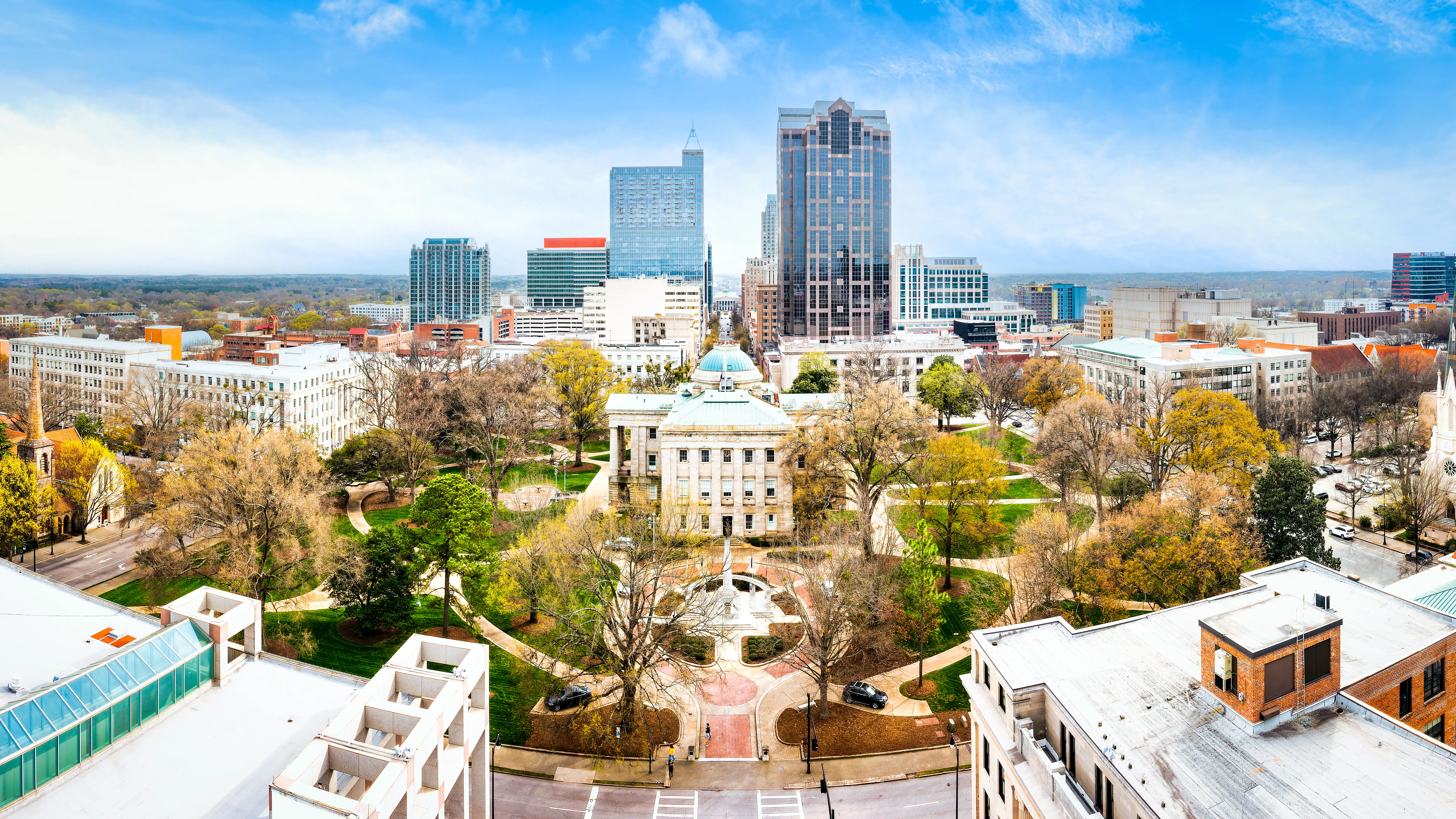 Downtown Raleigh skyline rises behind the North Carolina State Capitol and surrounding tree-filled grounds.