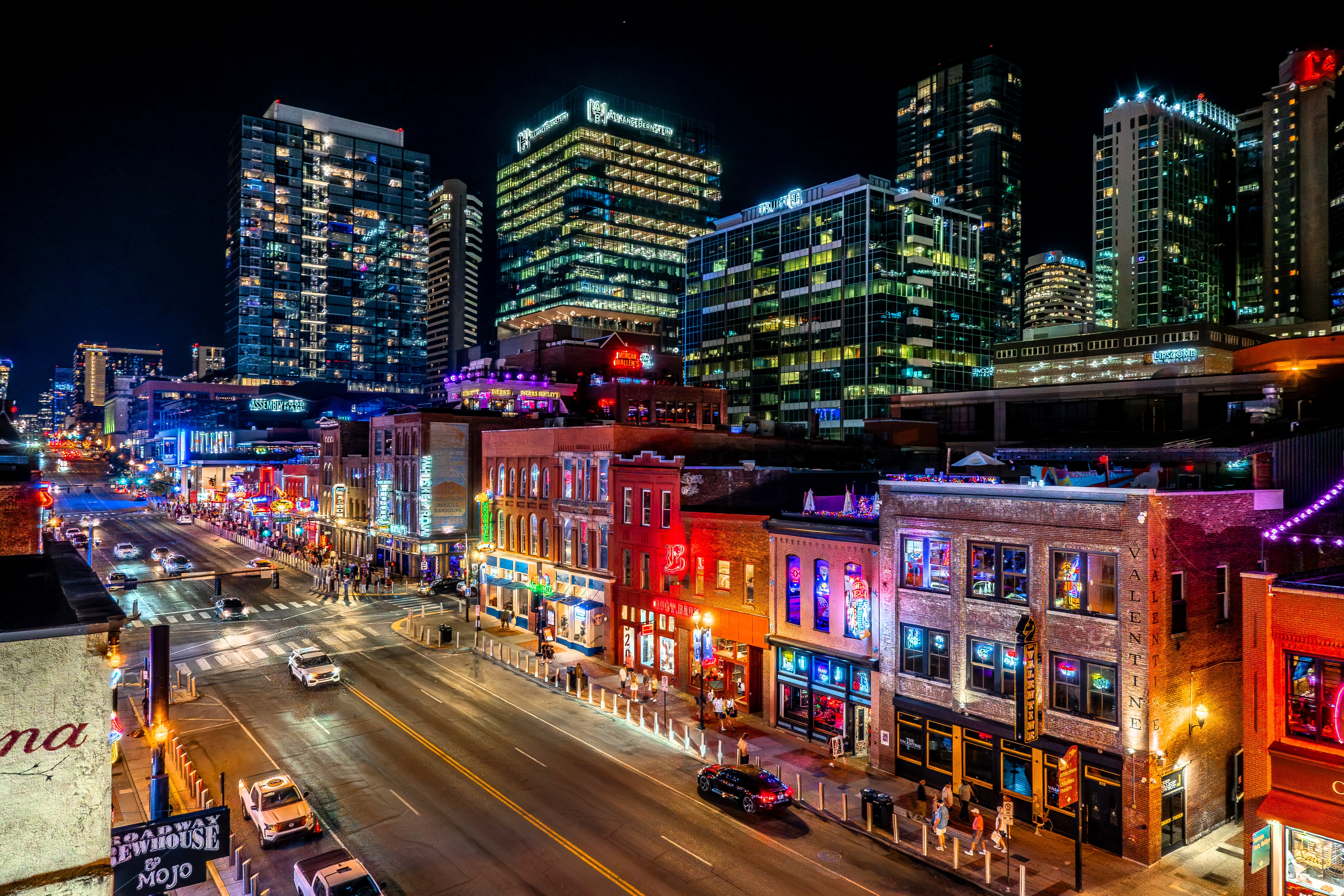 Neon-lit Broadway street in downtown Nashville with traffic, bars, and illuminated high-rise buildings at night.