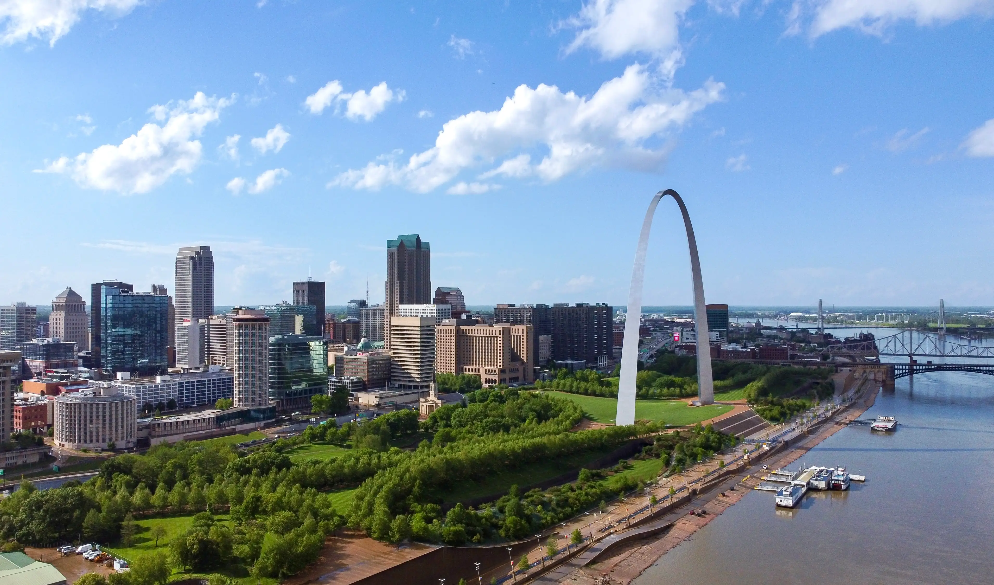 St. Louis skyline with the Gateway Arch beside a wide river under a partly cloudy blue sky.