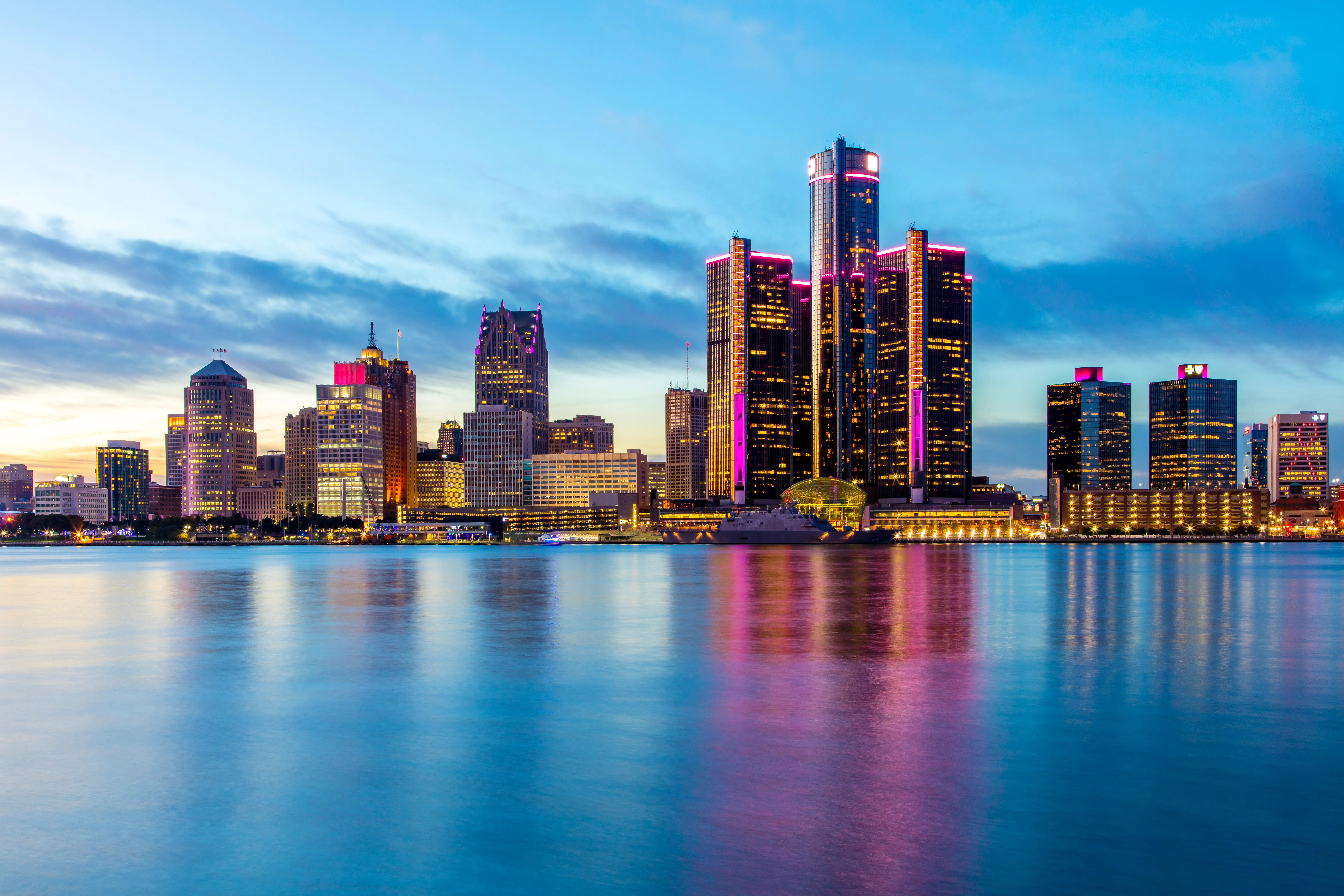Detroit skyline at dusk with the Renaissance Center illuminated and reflected across the river.