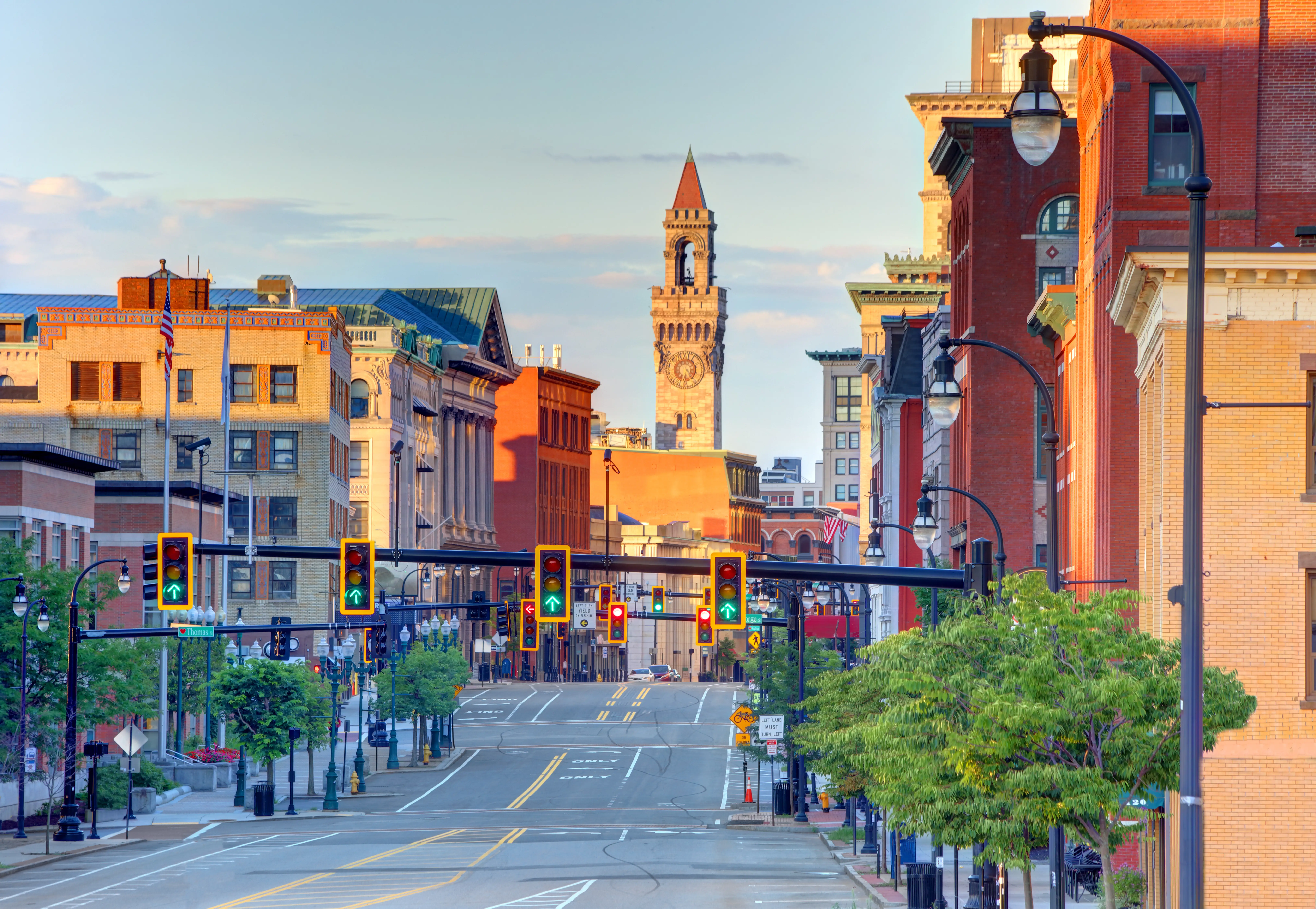 A quiet downtown street lined with brick buildings and traffic lights leads toward a clock tower at sunset.