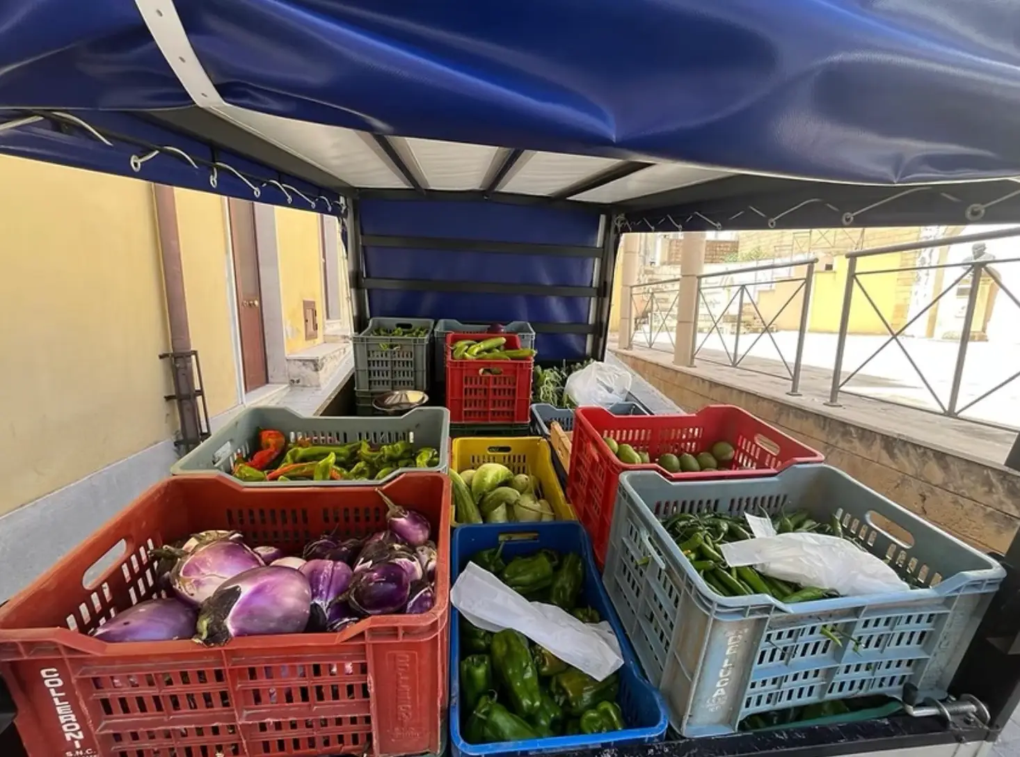 Plastic crates of fresh vegetables are arranged under a blue canopy on a market delivery vehicle.