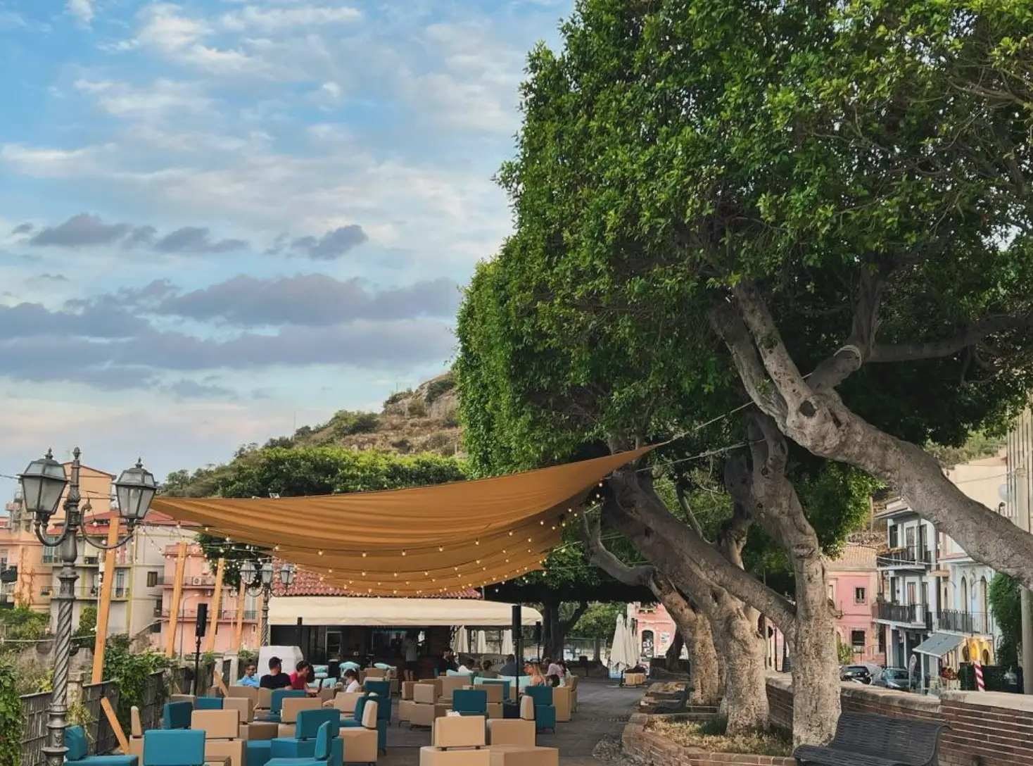 Outdoor café seating sits beneath a tan canopy and string lights beside large trees in a plaza in Melilli, where the author lives.