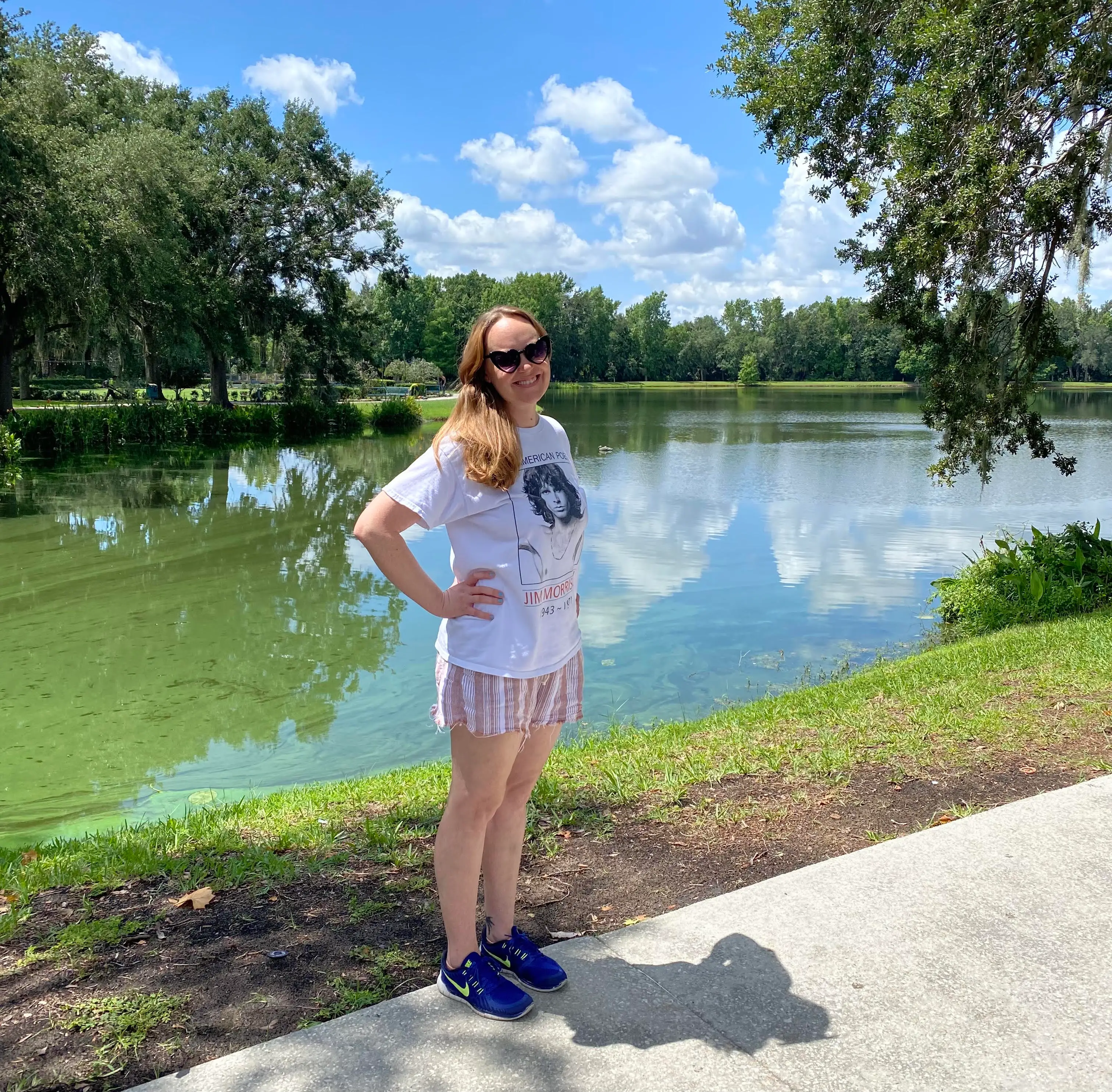 Person wearing sunglasses stands by a green lake with trees and cloud reflections.