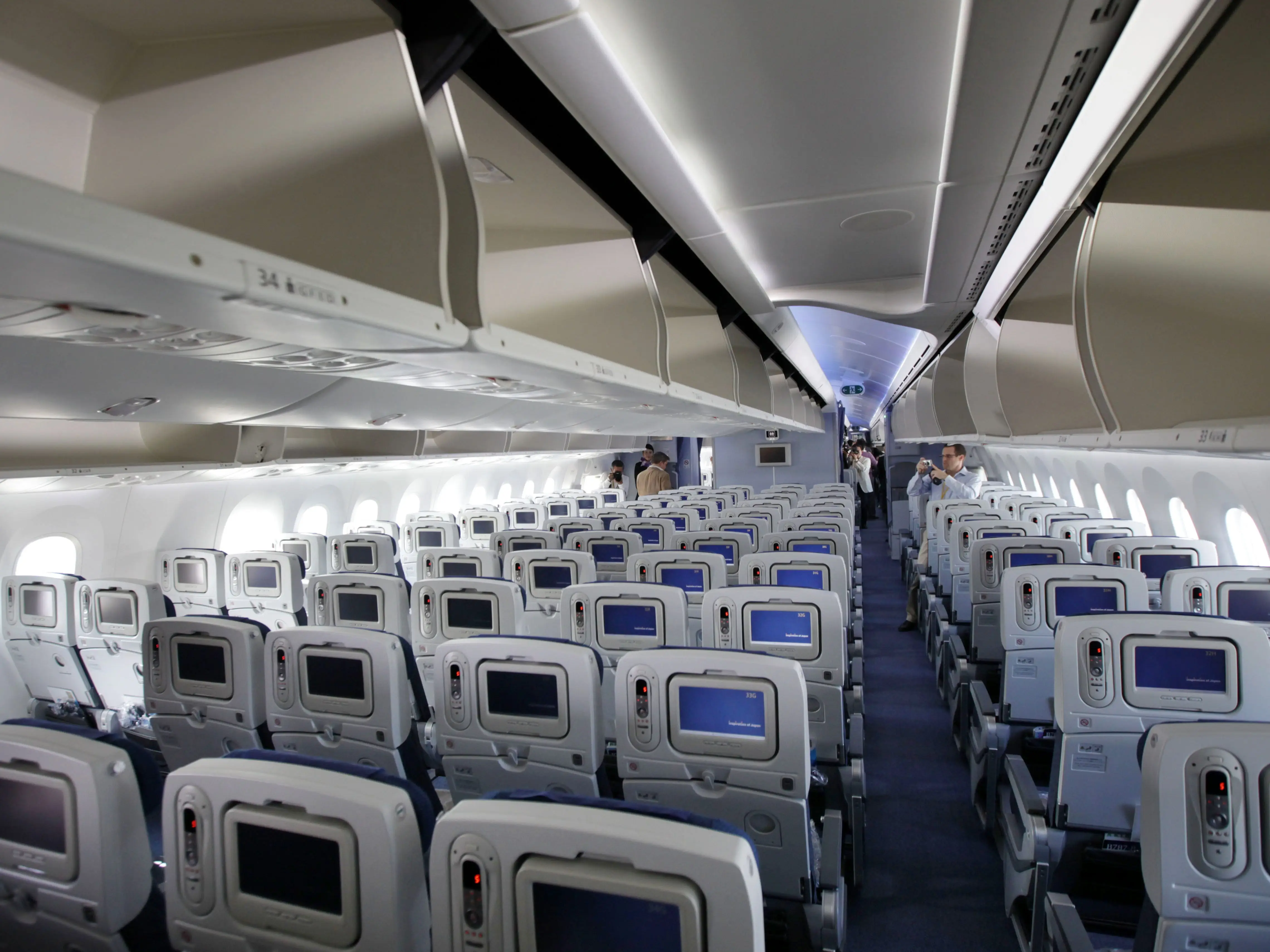 Economy-class Boeing 787 Dreamliner airplane cabin with rows of seatback screens, overhead bins, and a central aisle.