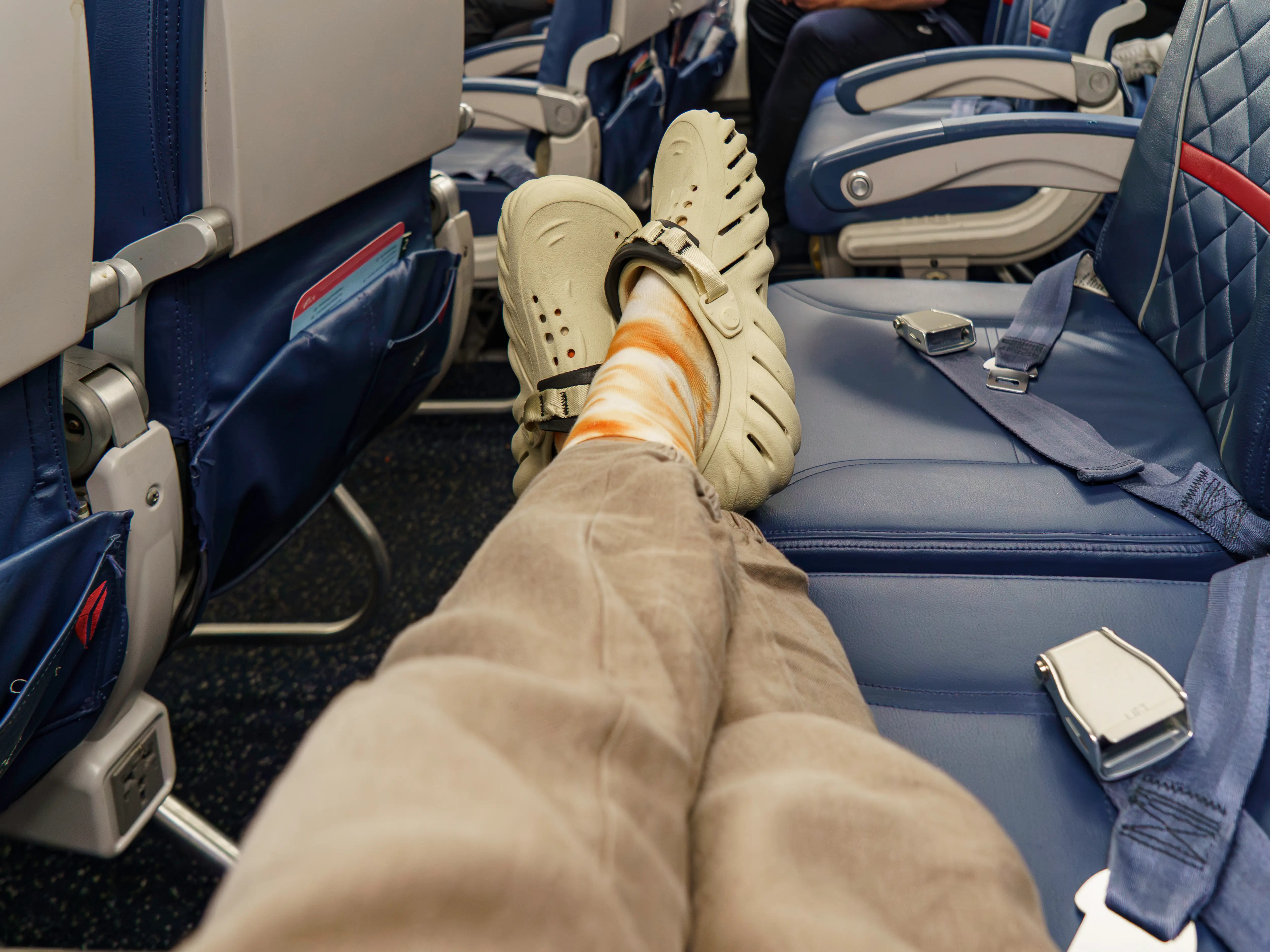 Passenger's feet in beige clogs rest on a blue airplane seat beside visible seat belts in the cabin.