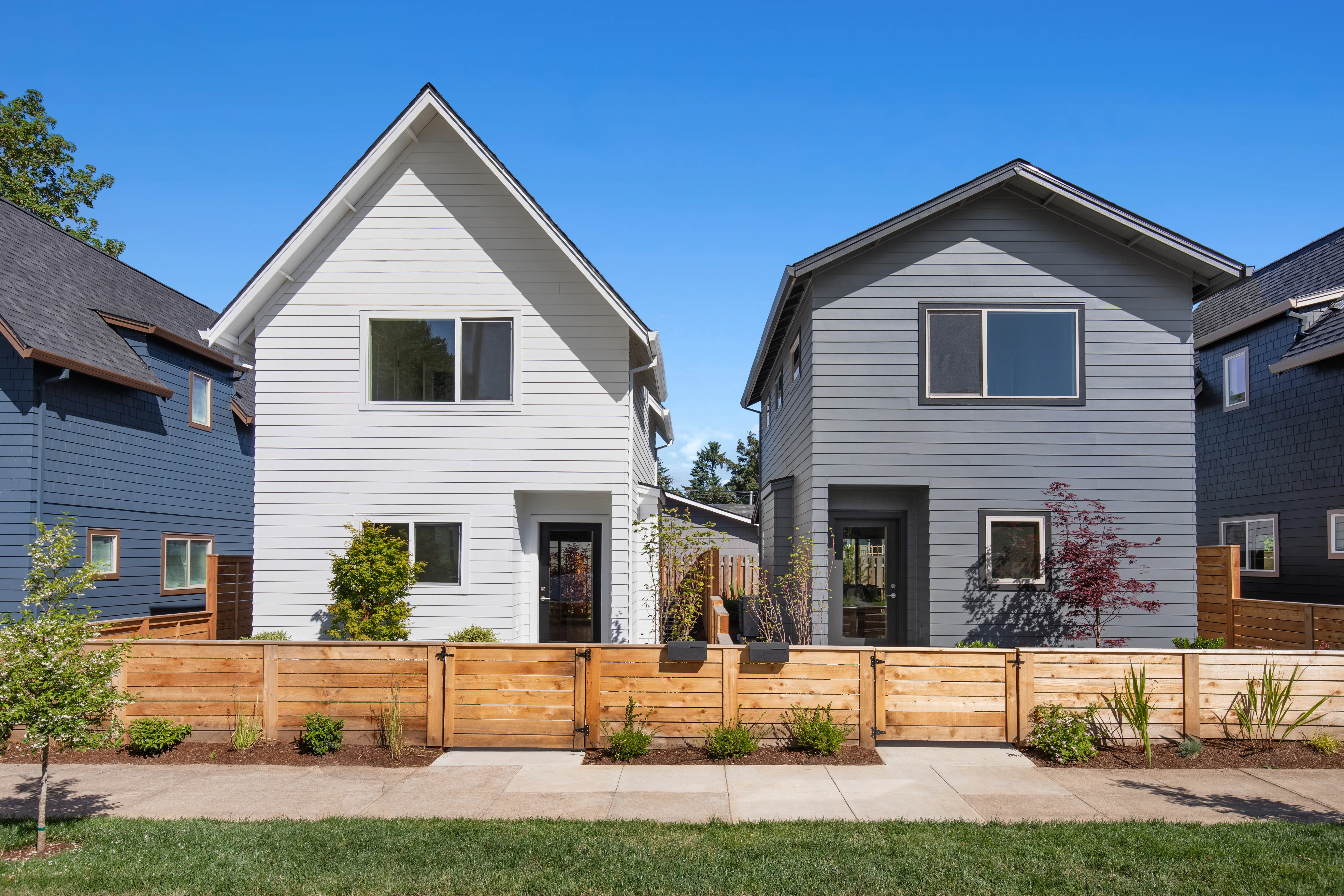 Two modern detached houses stand behind a wooden fence on a sunny residential street.