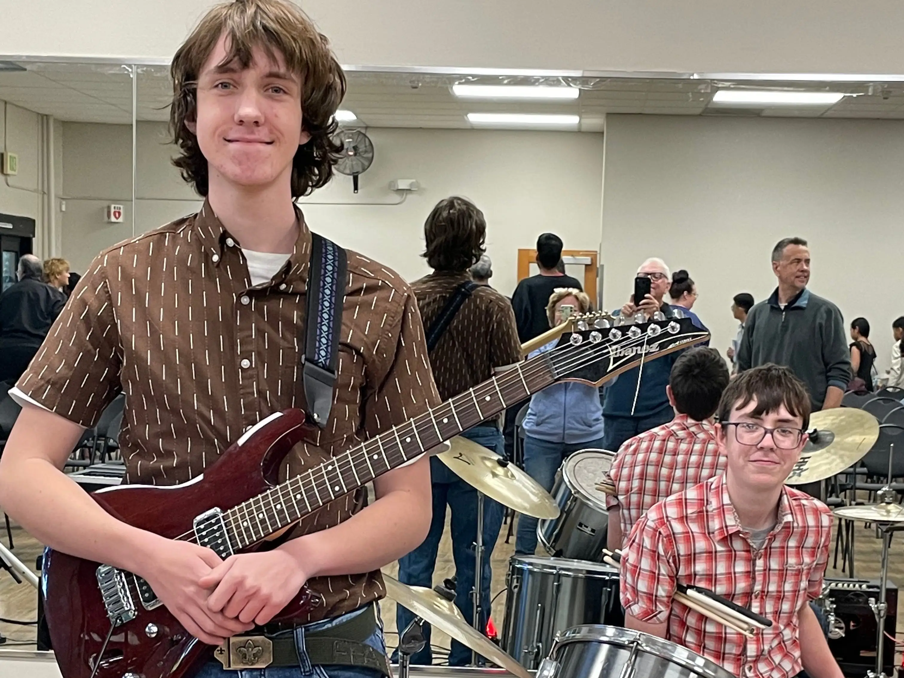 Two musicians pose with an electric guitar and drum kit in a mirrored rehearsal room.